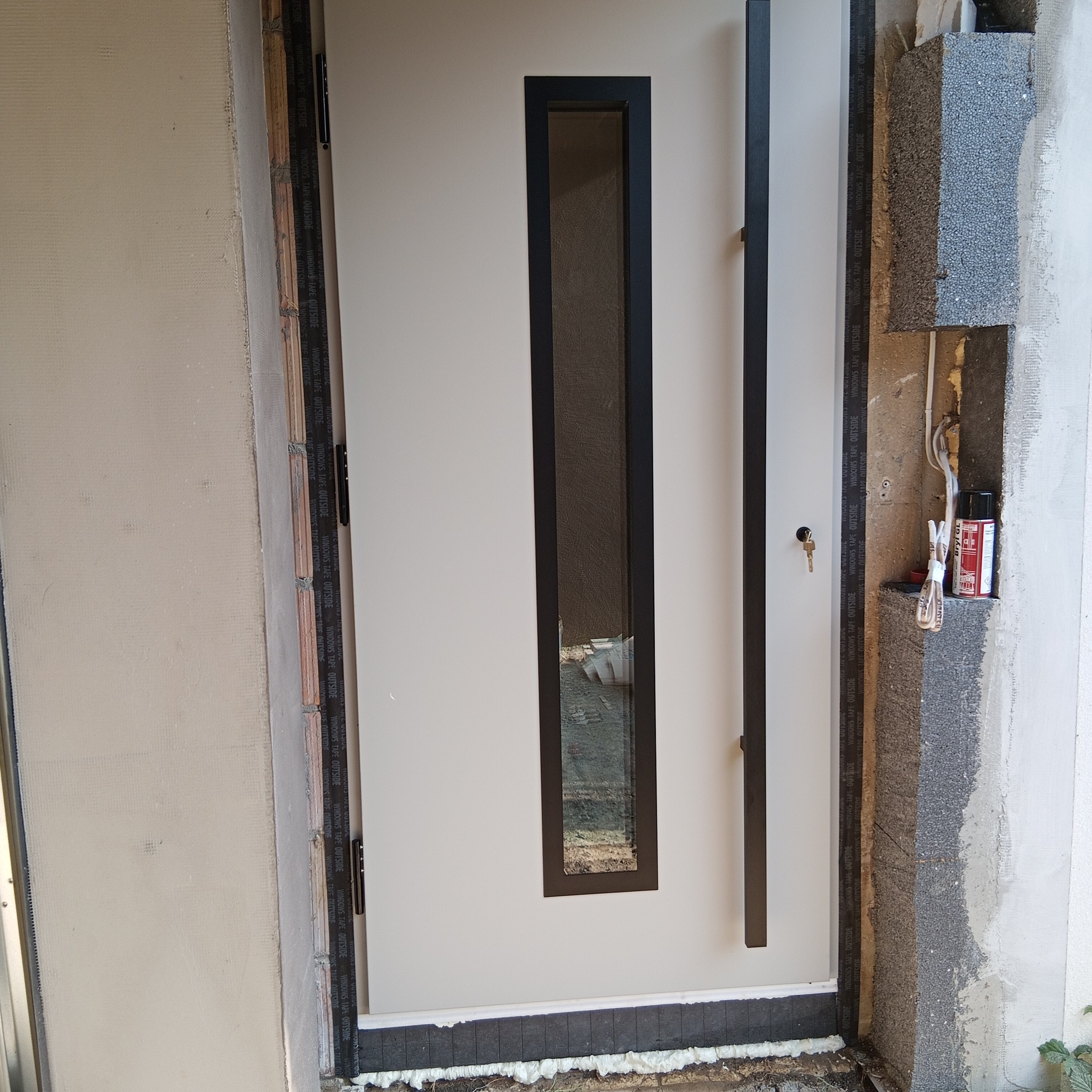 Białe drzwi wejściowe z czarną szybą i długim uchwytem, widoczne detale montażu z użyciem taśmy uszczelniającej i pianki montażowej. Klucz w zamku.