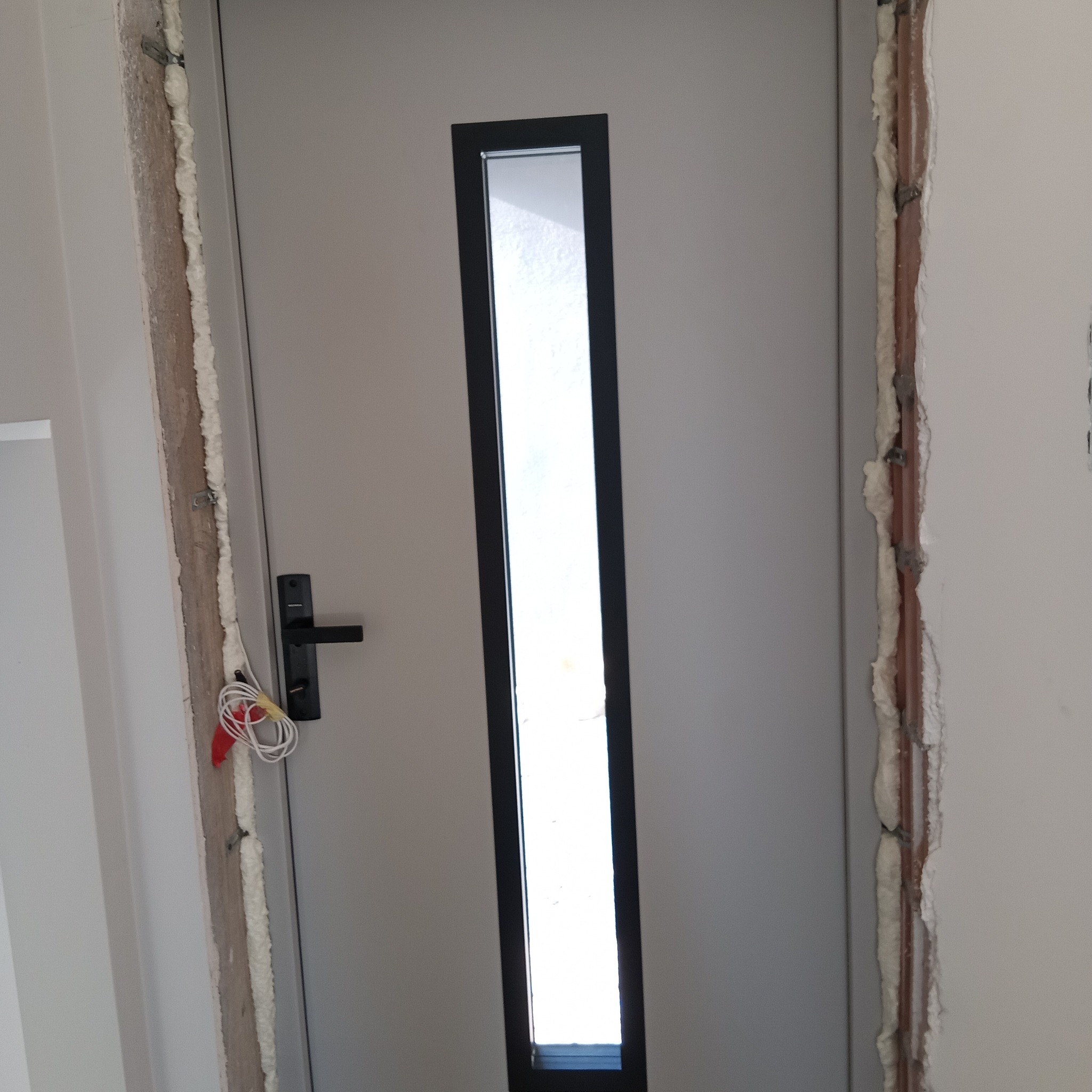 Szare drzwi z długim, wąskim przeszkleniem, osadzone w ościeżnicy z widoczną pianką montażową. Widoczne przewody elektryczne przy klamce. Ujęcie z bliska.