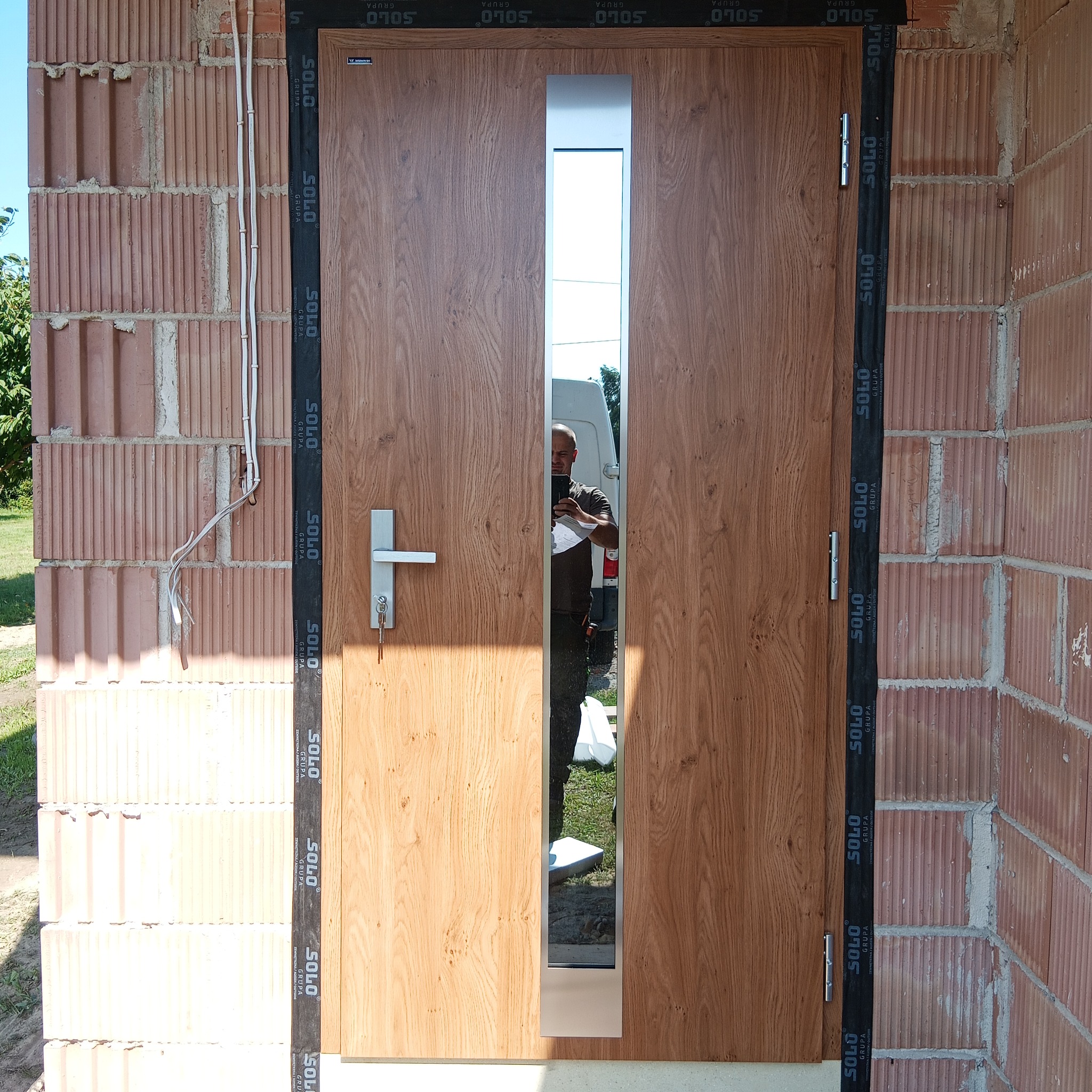 Zamontowane drzwi wejściowe z przeszkleniem, widoczne na tle surowej ściany z cegły. Na drzwiach widoczne oznaczenia producenta. W odbiciu szyby widać osobę robiącą zdjęcie.