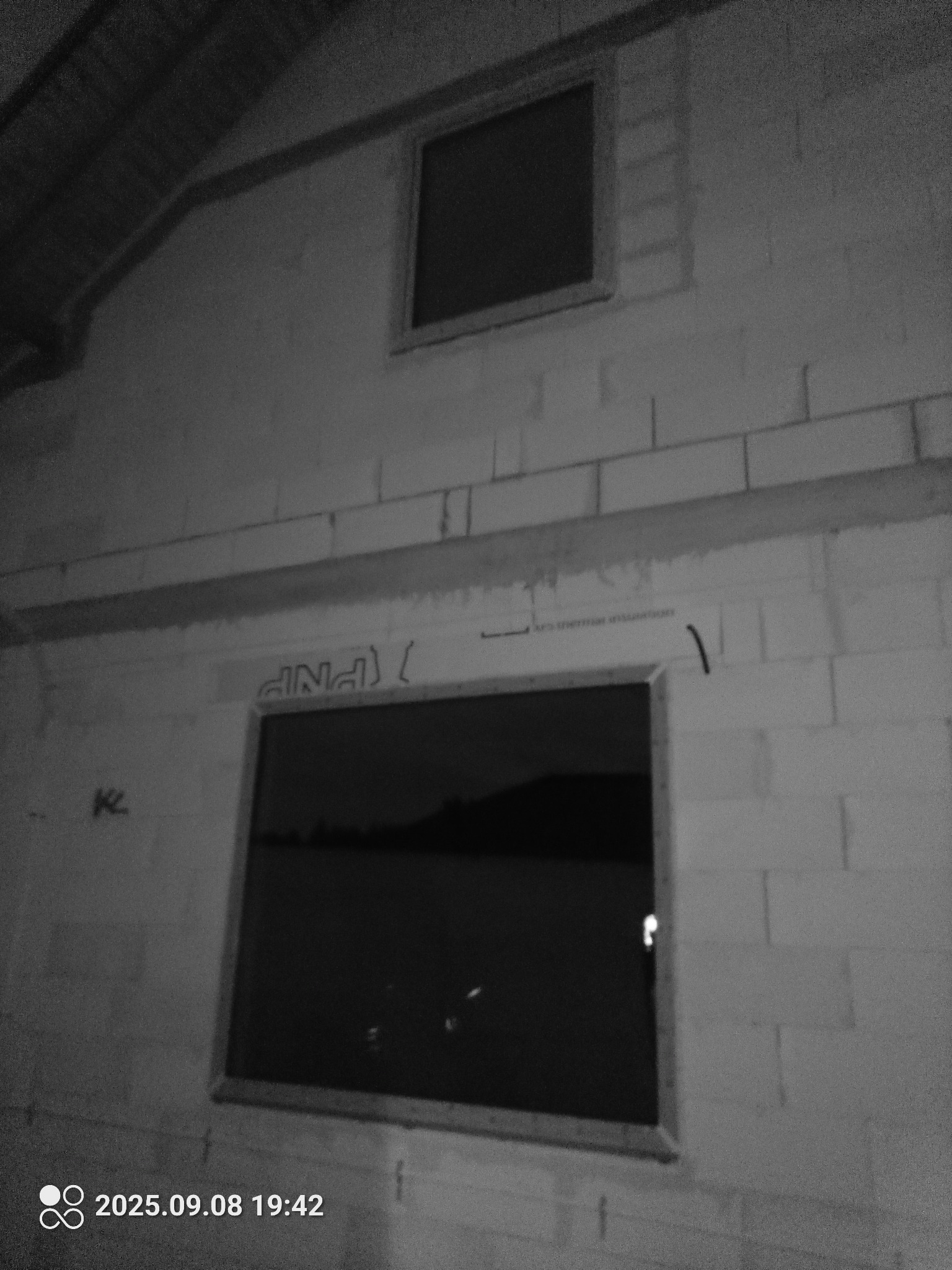 Surowy mur z dwoma oknami o różnych rozmiarach, w trakcie montażu. Widoczne niedoskonałości i brak wykończenia. Zdjęcie w odcieniach szarości, z datą w lewym dolnym rogu.