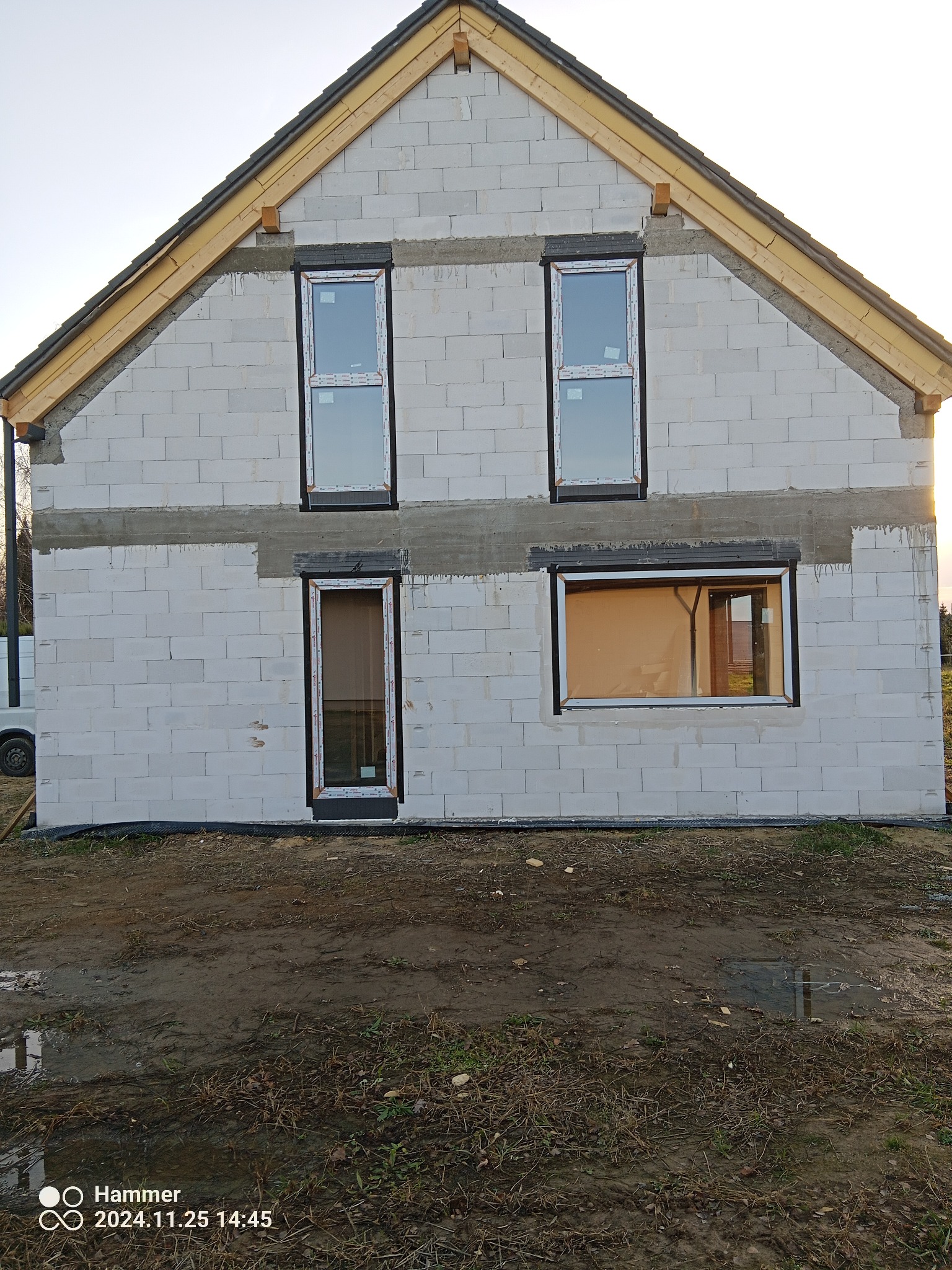 Surowy stan budynku z zamontowanymi oknami PCV o białych ramach. Widoczne bloczki betonowe, drewniana konstrukcja dachu i błotniste podłoże. Częściowo widoczny samochód dostawczy.