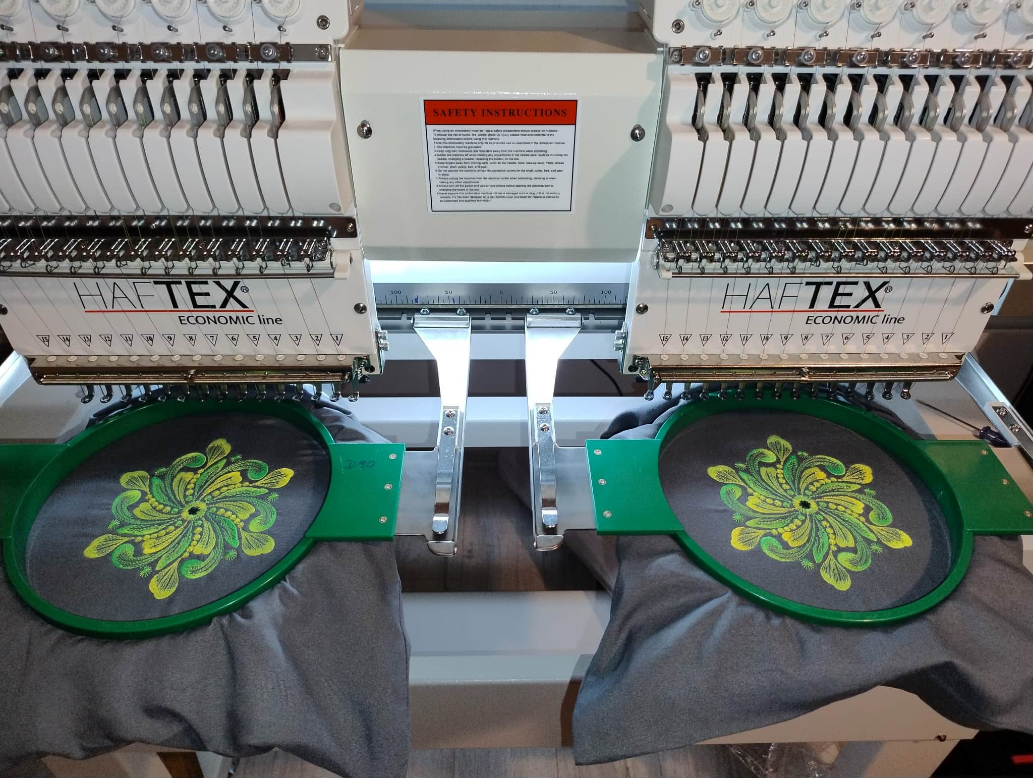 Automat do haftowania Haftex z dwoma głowicami, prezentujący wzór kwiatowy w odcieniach zieleni i żółci na szarej tkaninie, umieszczonej w zielonych tamborkach.