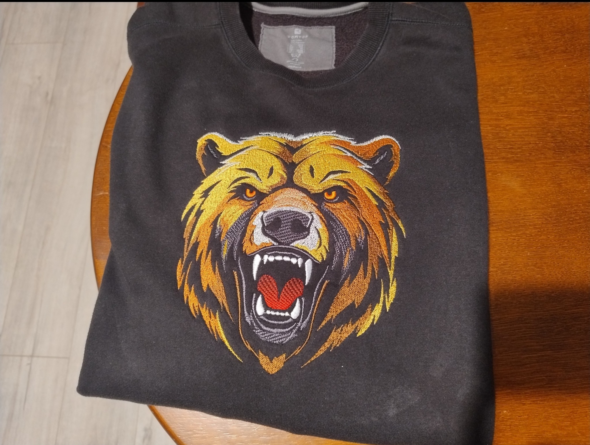 Ciemnoszara bluza z haftowanym wizerunkiem głowy niedźwiedzia z otwartą paszczą, leżąca na drewnianym stole. Widoczny fragment metki na karku.