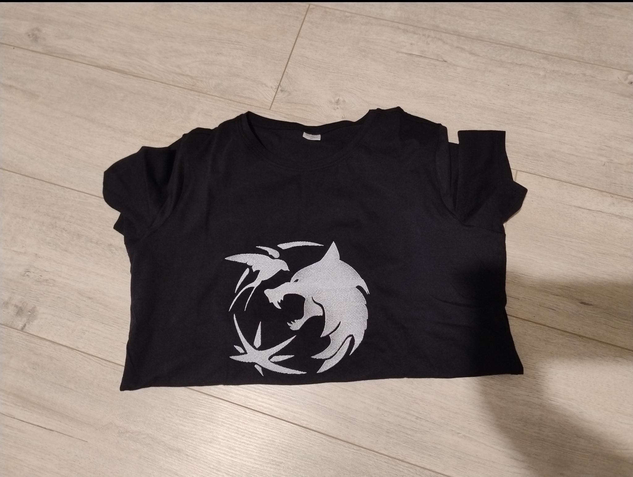 Czarna koszulka z szarym nadrukiem wilka na drewnianym tle. Grafika inspirowana motywem wiedźmińskim, leżąca płasko na jasnych panelach podłogowych.