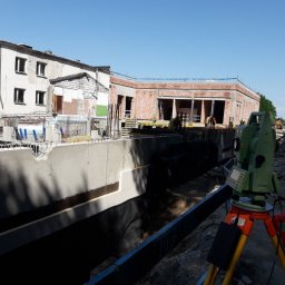 SG GEOLINE Sp. z o.o. - Zielony teodolit na statywie na tle budowy z fundamentami i budynkiem w tle. Prace pomiarowe w toku, widoczni pracownicy w kaskach. Słoneczny dzień.