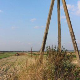 SG GEOLINE Sp. z o.o. - Betonowy trójnóg geodezyjny na tle pola uprawnego z pracującym traktorem w oddali, widok z dołu, słoneczny dzień.