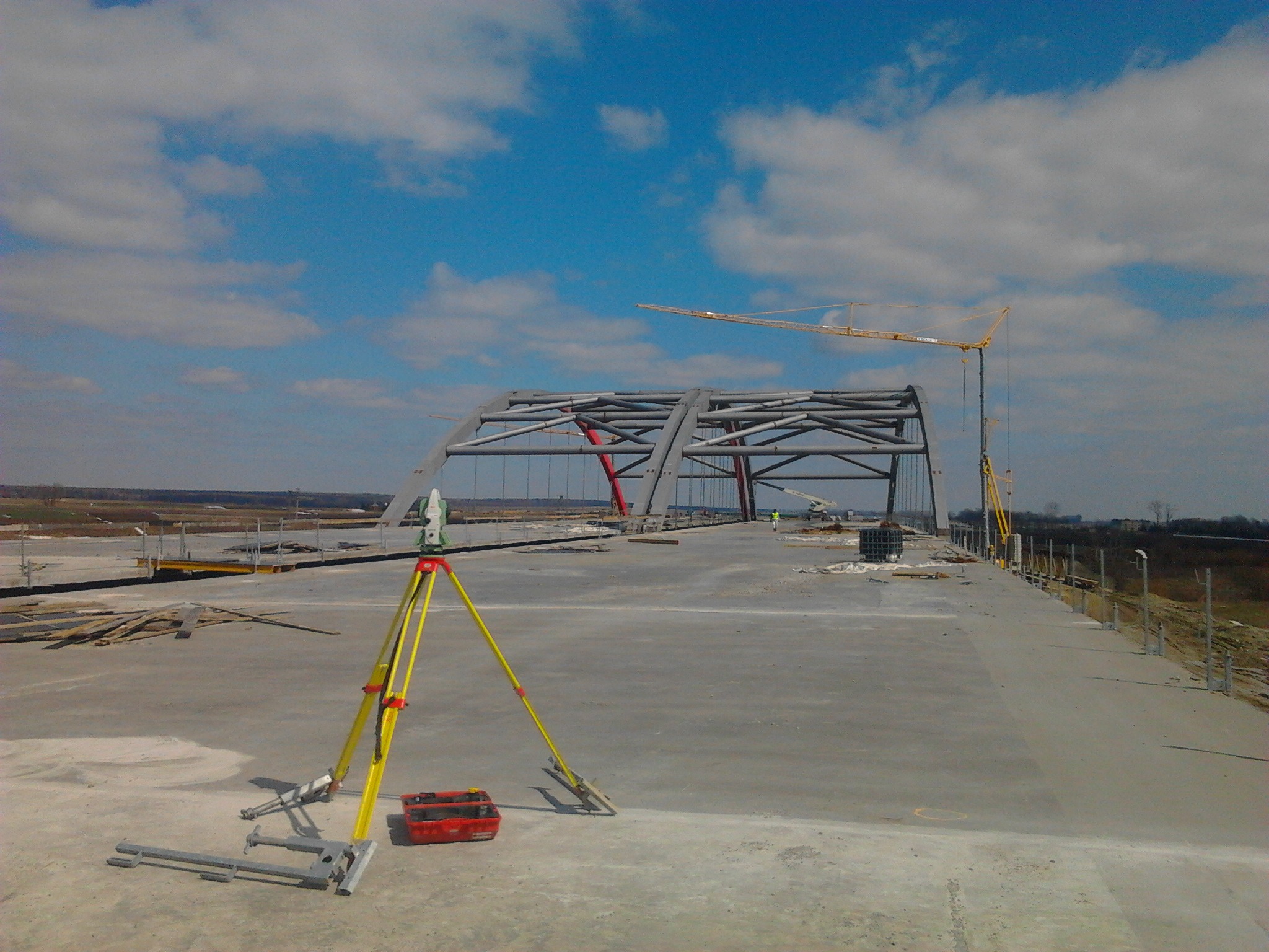 Prace pomiarowe na budowie mostu: żółty teodolit na tle stalowej konstrukcji łukowej i dźwigu, błękitne niebo z chmurami w tle. Widoczne narzędzia i elementy konstrukcyjne.