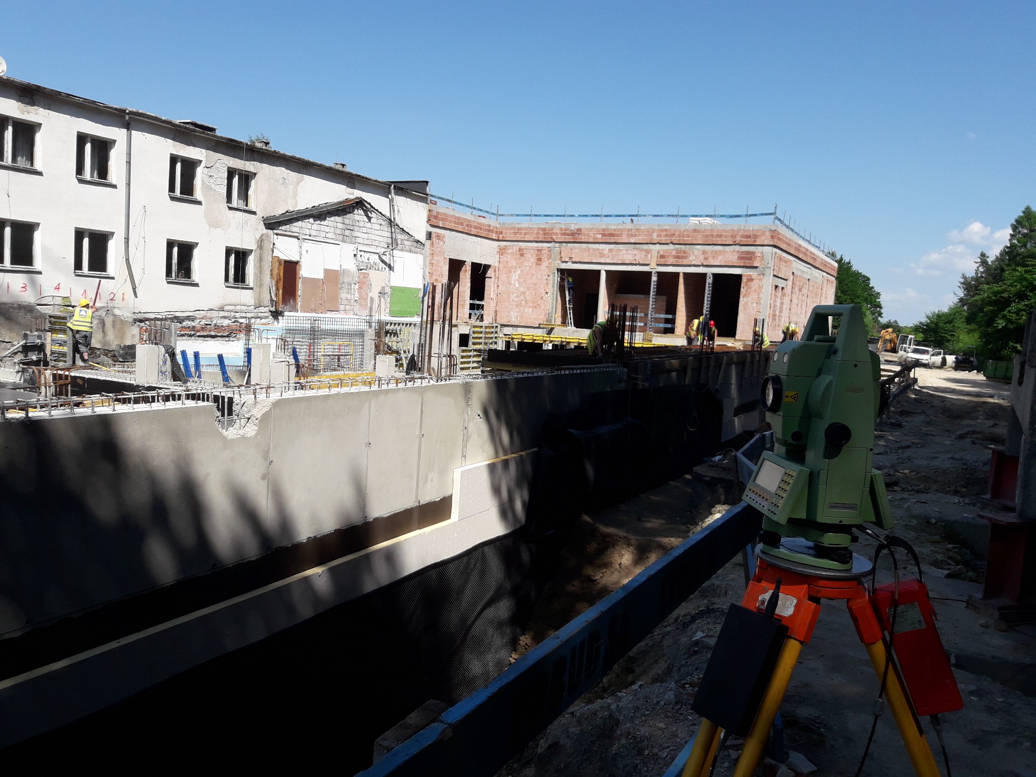 Zielony teodolit na statywie na tle budowy z fundamentami i budynkiem w tle. Prace pomiarowe w toku, widoczni pracownicy w kaskach. Słoneczny dzień.