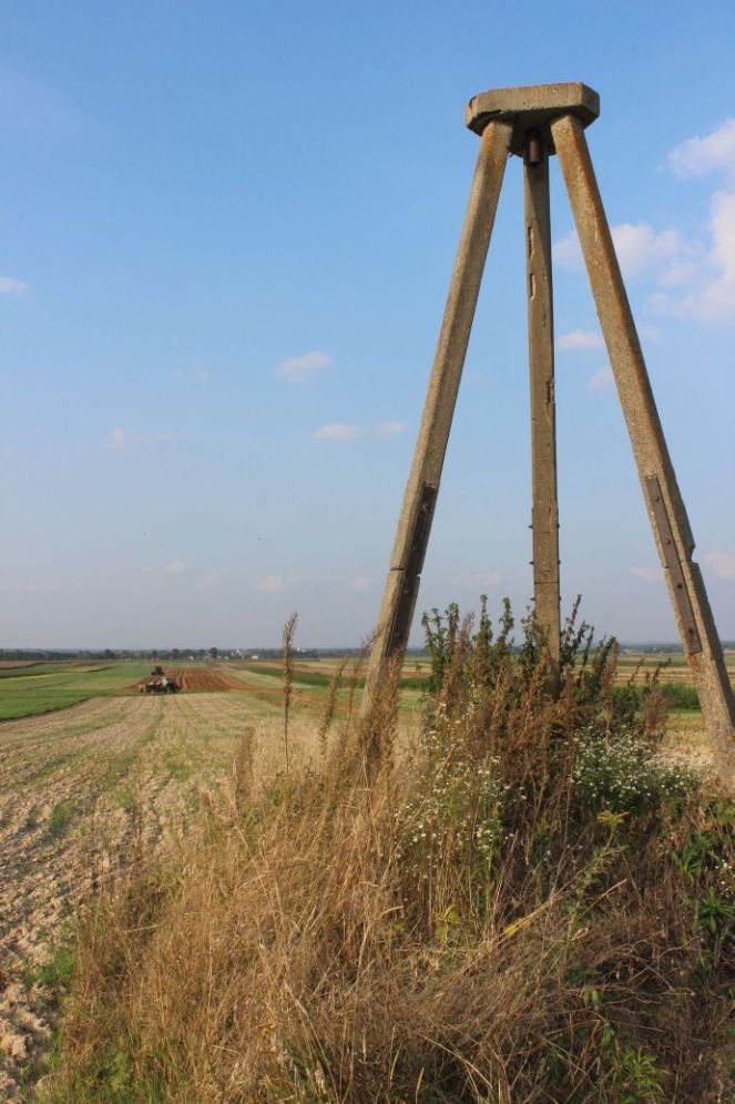 Betonowy trójnóg geodezyjny na tle pola uprawnego z pracującym traktorem w oddali, widok z dołu, słoneczny dzień.