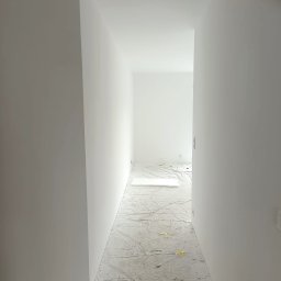 Magdalena Kruszewska - Białe, świeżo pomalowane wnętrze korytarza z zabezpieczoną folią podłogą. Widok w głąb pomieszczenia, minimalistyczny styl, surowy stan po malowaniu.