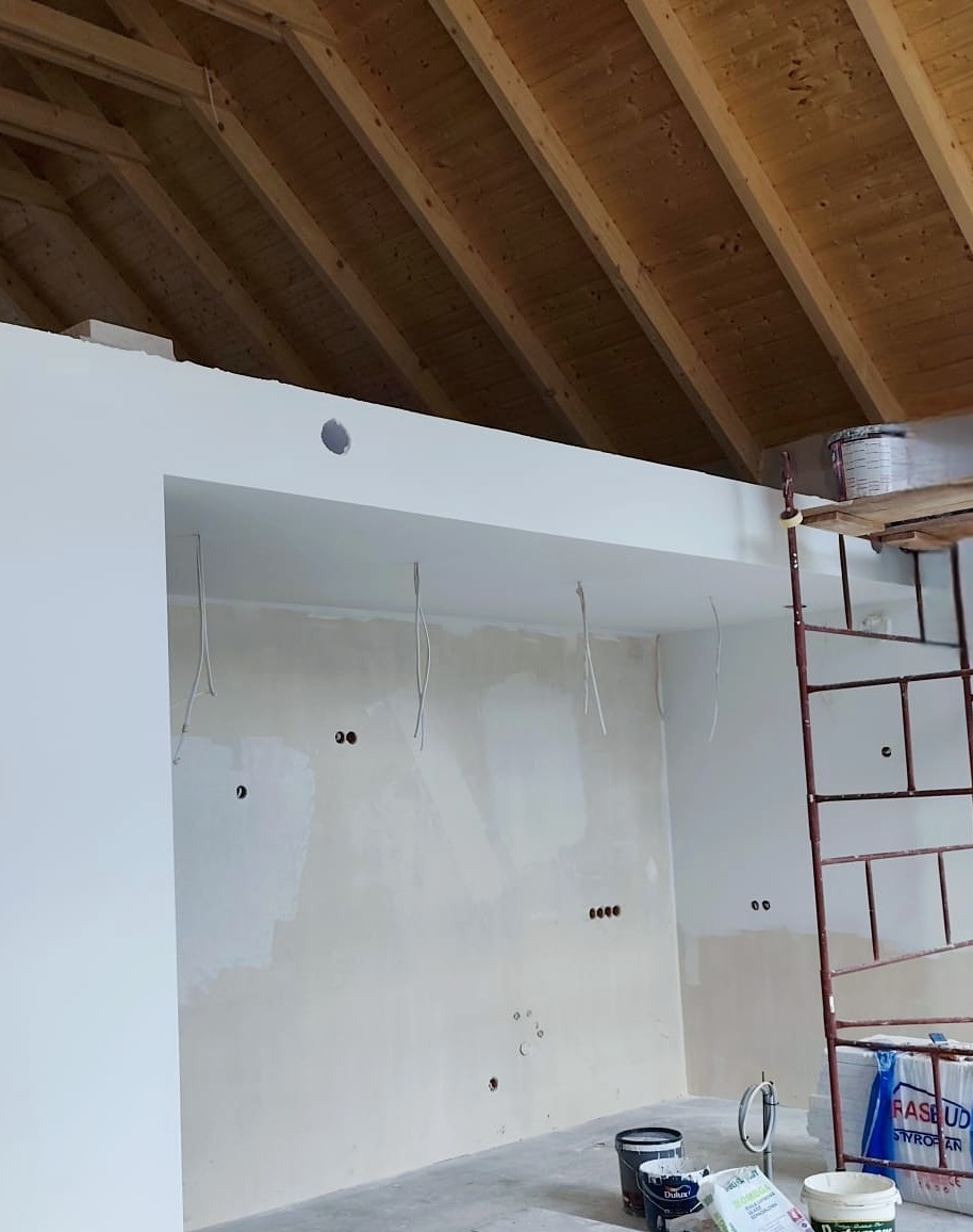 Wnętrze w trakcie prac wykończeniowych: szpachlowana ściana z widocznymi przewodami, drewniany strop, rusztowanie i farby. Etap przygotowania do malowania.