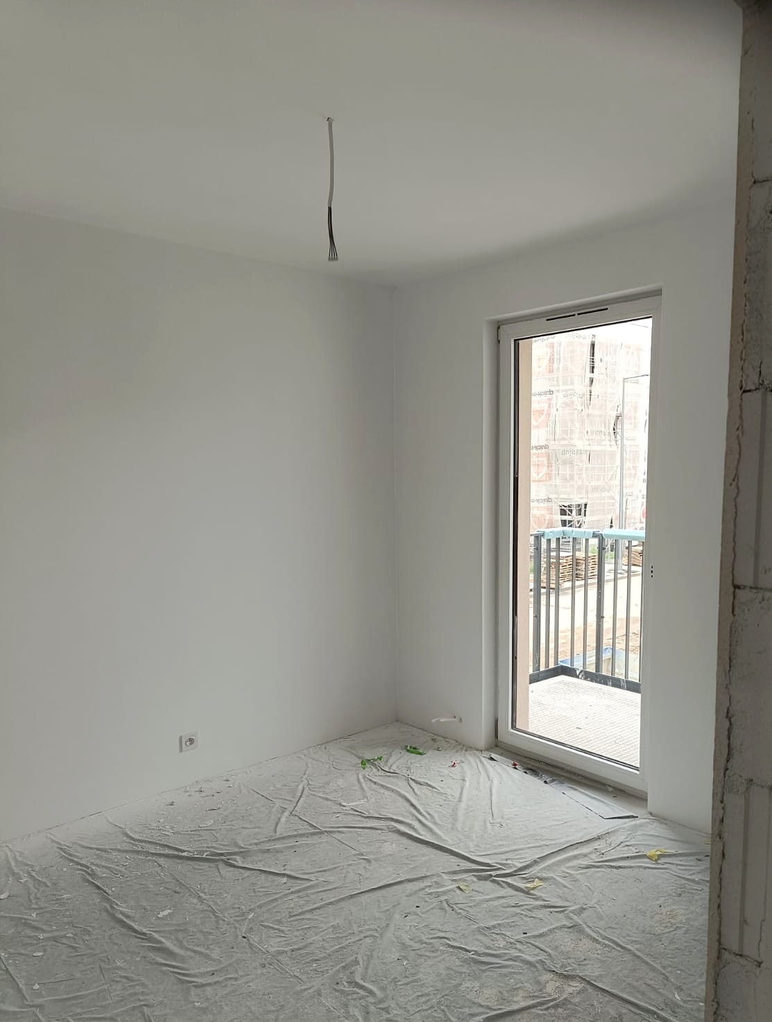 Białe ściany w pokoju po malowaniu, zabezpieczona podłoga folią malarską, widoczne kable wystające z sufitu i okno balkonowe. Świeżo pomalowane wnętrze.