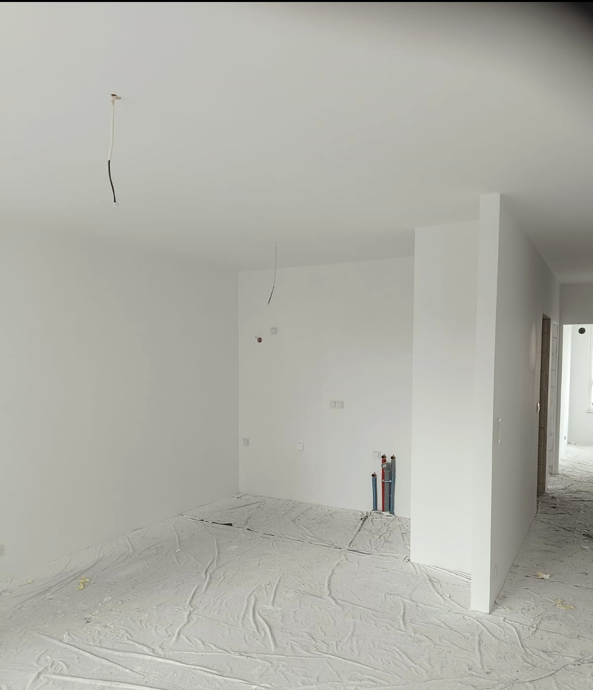 Białe, świeżo pomalowane wnętrze w trakcie remontu. Podłoga zabezpieczona folią malarską, widoczne wystające kable i rury. Minimalistyczny styl, jasne oświetlenie.