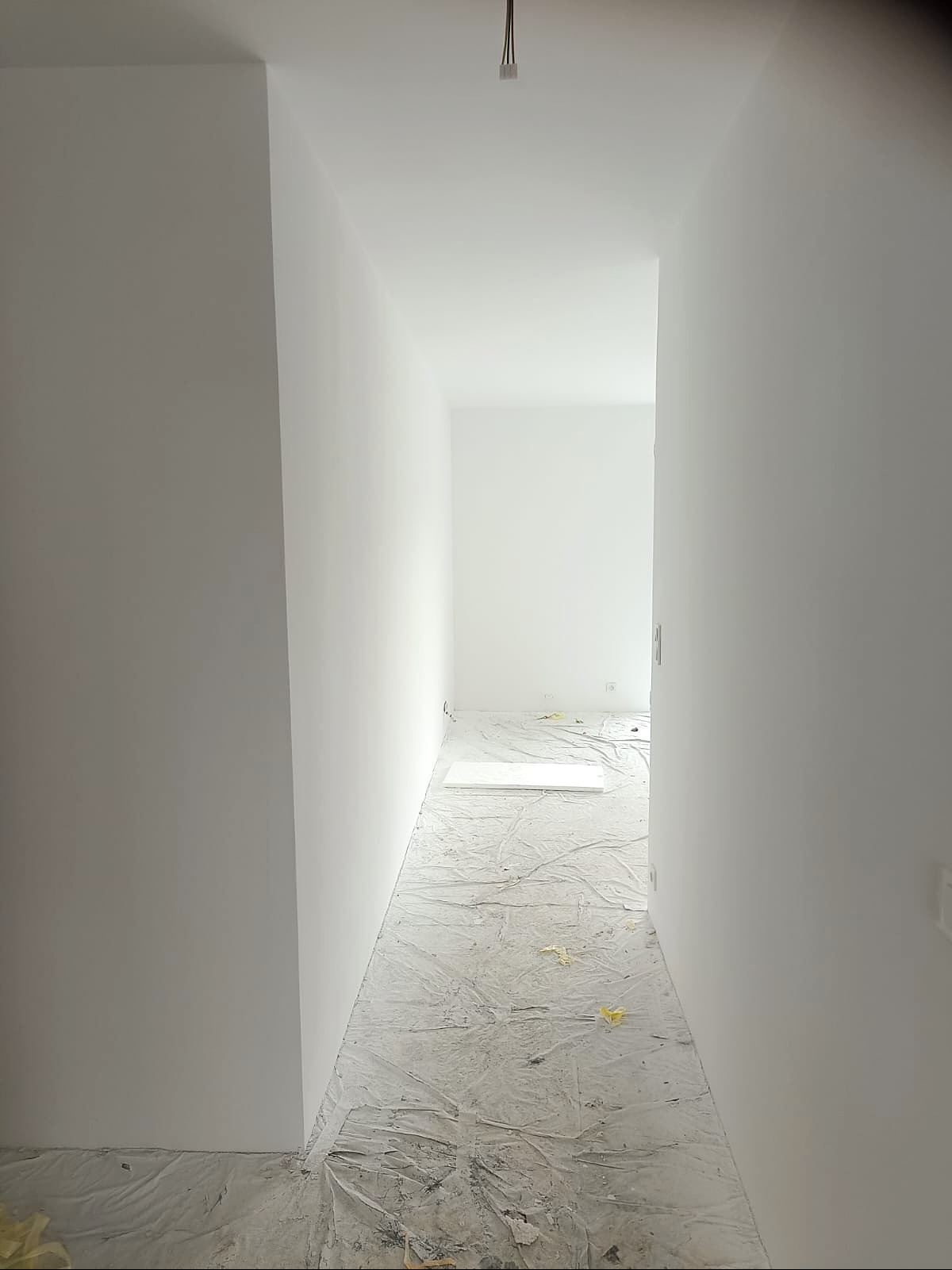 Białe, świeżo pomalowane wnętrze korytarza z zabezpieczoną folią podłogą. Widok w głąb pomieszczenia, minimalistyczny styl, surowy stan po malowaniu.