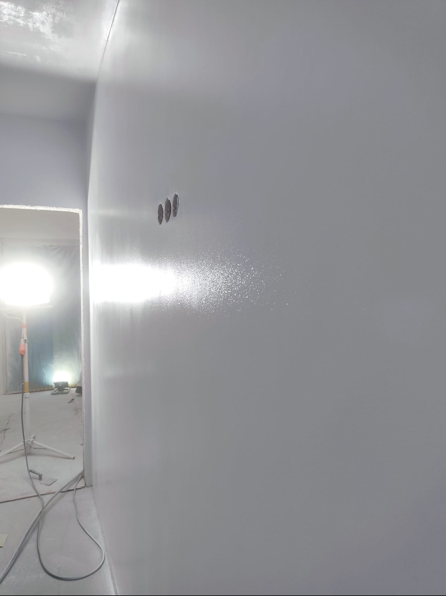 Świeżo pomalowana, gładka ściana w odcieniach szarości z trzema otworami na gniazdka. W tle widoczne oświetlenie budowlane i fragment pomieszczenia w trakcie prac wykończeniowych.