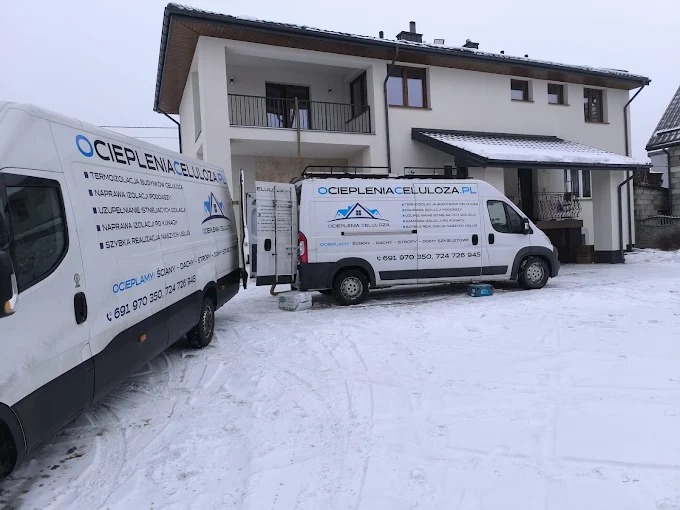 Dwa białe busy z logo firmy ocieplającej celulozą na tle ośnieżonego domu z jasną elewacją. Jeden bus z otwartymi drzwiami. Zima, pochmurne niebo.