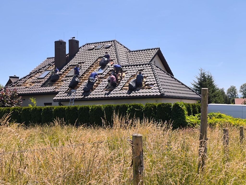 Ekipa montuje izolację na dachu domu jednorodzinnego z ciemną dachówką, widoczne okna dachowe i komin. Prace w toku, widok zza wysokiej trawy.