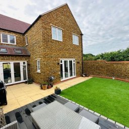 Dwupiętrowy dom z kamienia z ogrodem i tarasem. Widoczne białe okna i drzwi balkonowe, trawnik z kamiennym murem. Grill i meble ogrodowe na pierwszym planie.