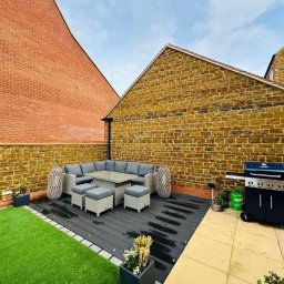 Umeblowany taras z szarą sofą narożną, stołem i pufami na ciemnej desce kompozytowej. Obok grill gazowy. Tło stanowią mury z cegły i kamienia.