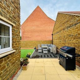 Ukończony taras z kostki brukowej i drewnianego podestu z meblami ogrodowymi oraz grillem, otoczony budynkami z cegły w ciepłych odcieniach.