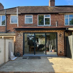 Nowoczesne, przesuwne drzwi tarasowe w ceglanym domu jednorodzinnym. Widoczne elementy wykończenia i przygotowany taras. Jasne, słoneczne światło.