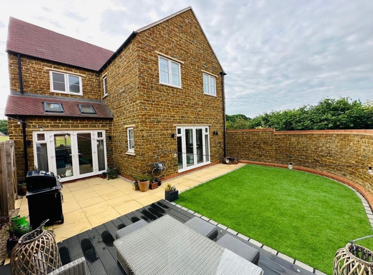 Dwupiętrowy dom z kamienia z ogrodem i tarasem. Widoczne białe okna i drzwi balkonowe, trawnik z kamiennym murem. Grill i meble ogrodowe na pierwszym planie.