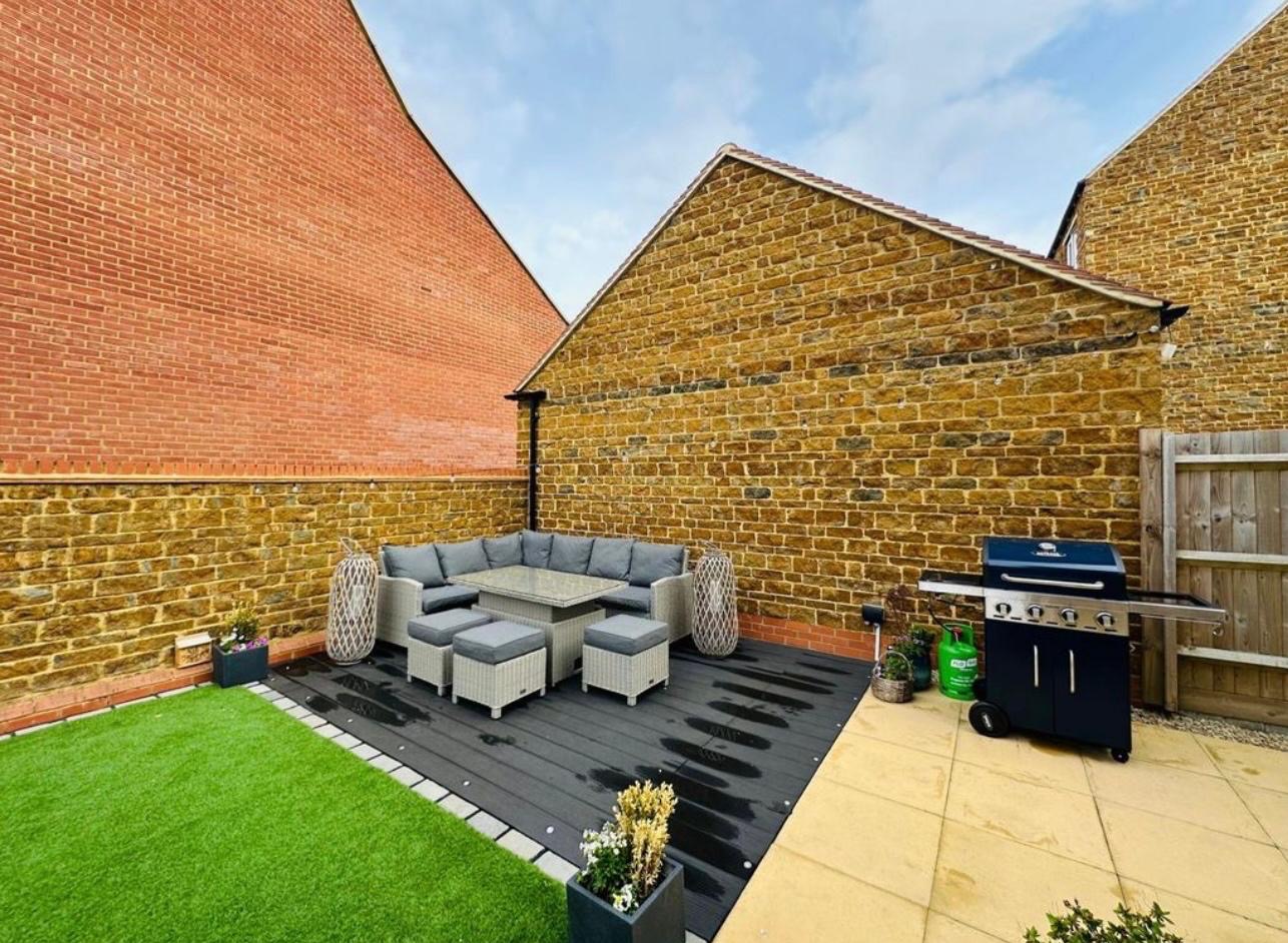 Umeblowany taras z szarą sofą narożną, stołem i pufami na ciemnej desce kompozytowej. Obok grill gazowy. Tło stanowią mury z cegły i kamienia.