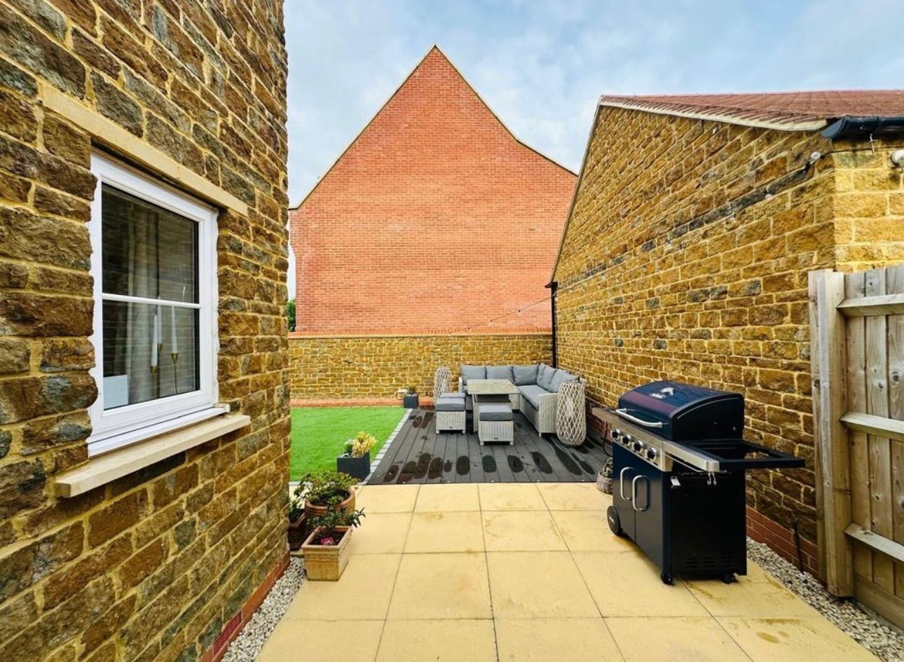 Ukończony taras z kostki brukowej i drewnianego podestu z meblami ogrodowymi oraz grillem, otoczony budynkami z cegły w ciepłych odcieniach.