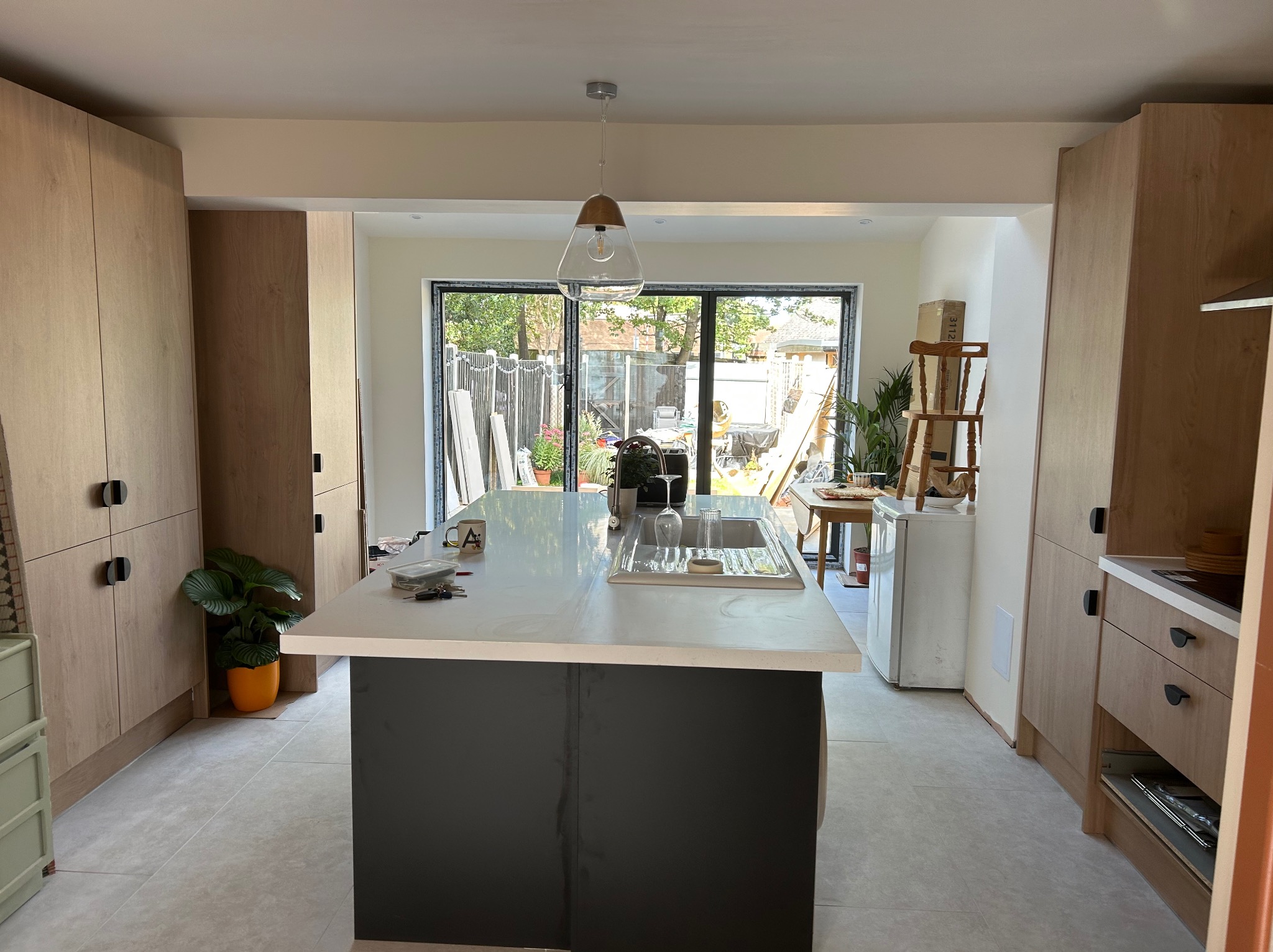 Jasna kuchnia z wyspą i drewnianymi szafkami, widok na ogród przez duże okno. Minimalistyczna lampa wisząca nad wyspą. Roślina doniczkowa dodaje akcentu.