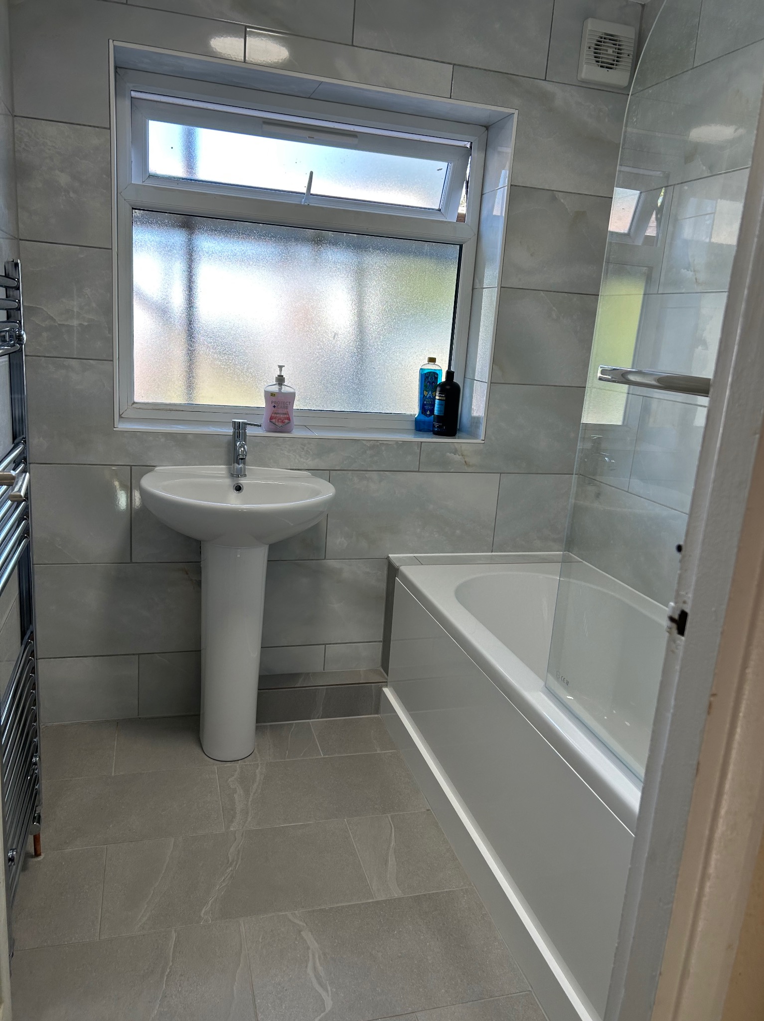 Jasna łazienka po remoncie: szare płytki imitujące marmur na ścianach i podłodze, biała umywalka na postumencie, wanna z parawanem i grzejnik drabinkowy.