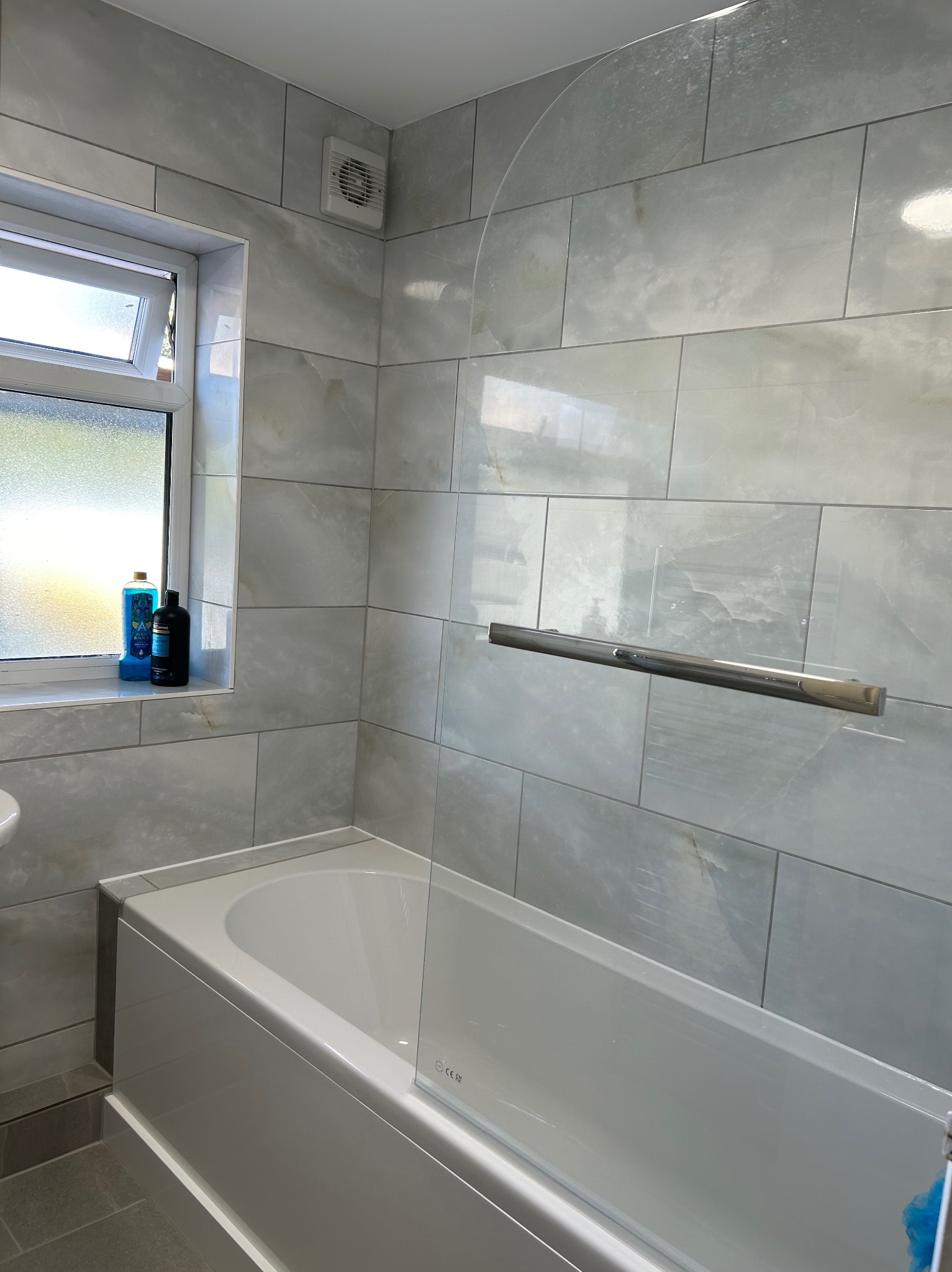 Wykończona łazienka z wanną, szklanym parawanem i płytkami imitującymi marmur. Na parapecie okiennym stoją kosmetyki. Widoczny wentylator.