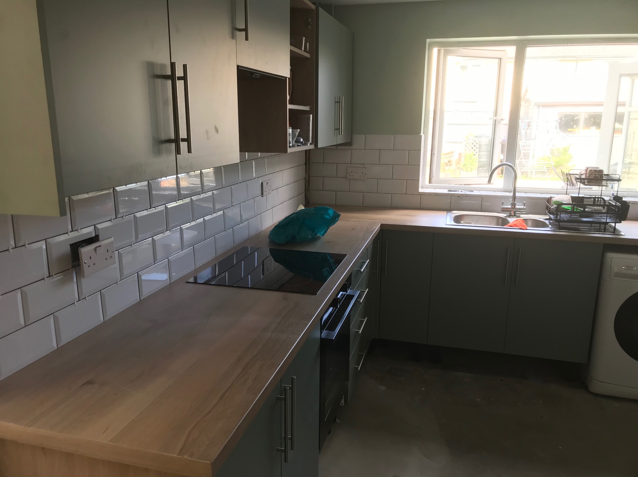 Jasna kuchnia z szarymi szafkami, drewnianym blatem i białymi płytkami. Widoczny zlew, płyta indukcyjna i pralka. Okno w tle wpuszcza dużo światła.