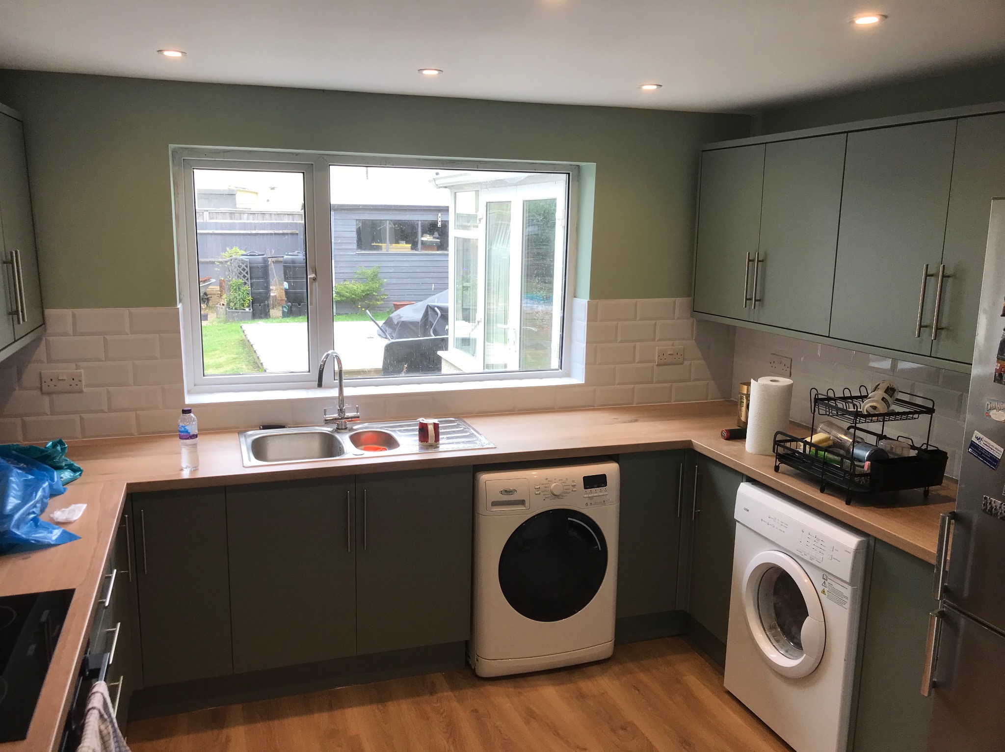 Jasna kuchnia z zielonymi szafkami, drewnianym blatem i białymi płytkami. Dwie pralki w zabudowie pod blatem. Widok na ogród przez okno.