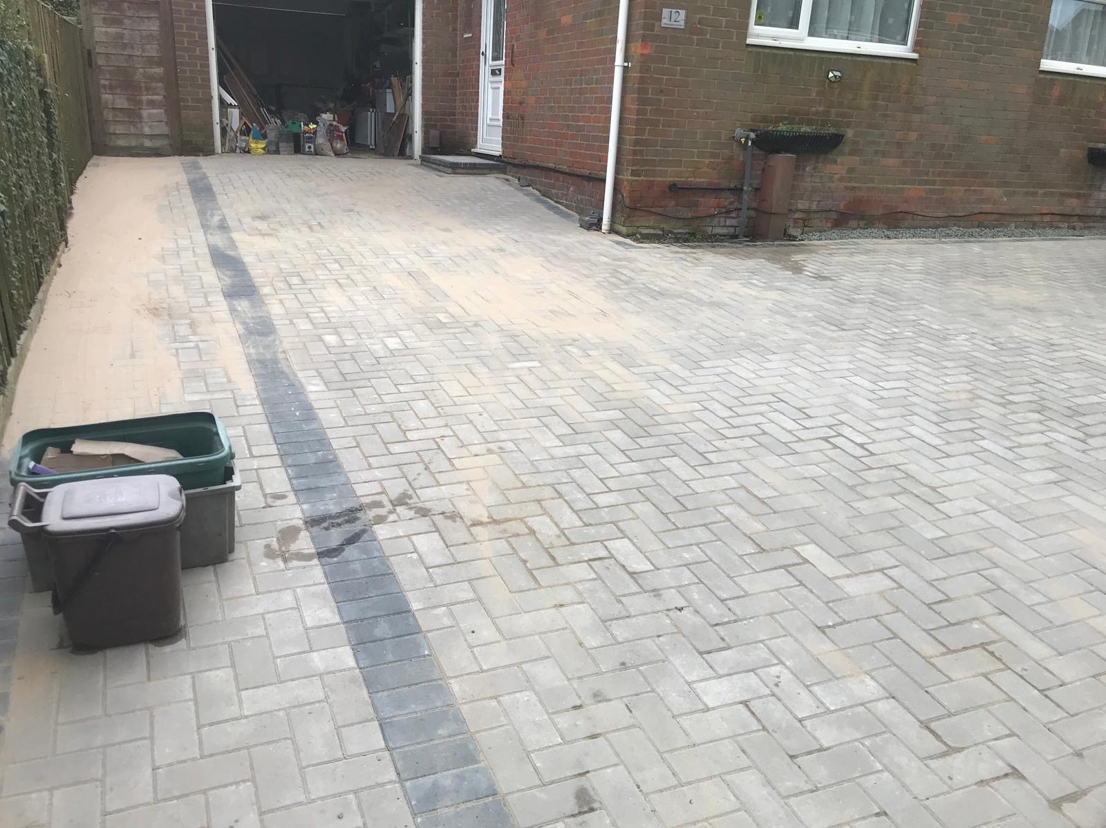 Ułożona kostka brukowa na podjeździe do domu z garażem, widoczne pojemniki na śmieci i piasek na obrzeżach. Ułożenie w jodełkę z ciemniejszym pasem.