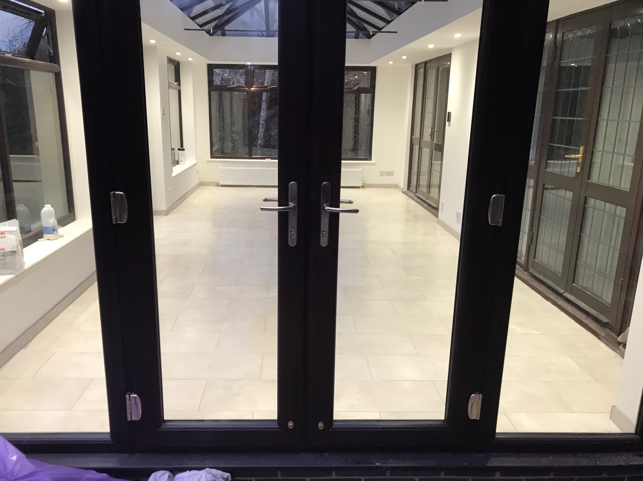 Jasne, przestronne wnętrze oranżerii z kremową podłogą z płytek, widoczne przez otwarte, ciemne, dwuskrzydłowe drzwi. Okna z ciemnymi ramami w tle.