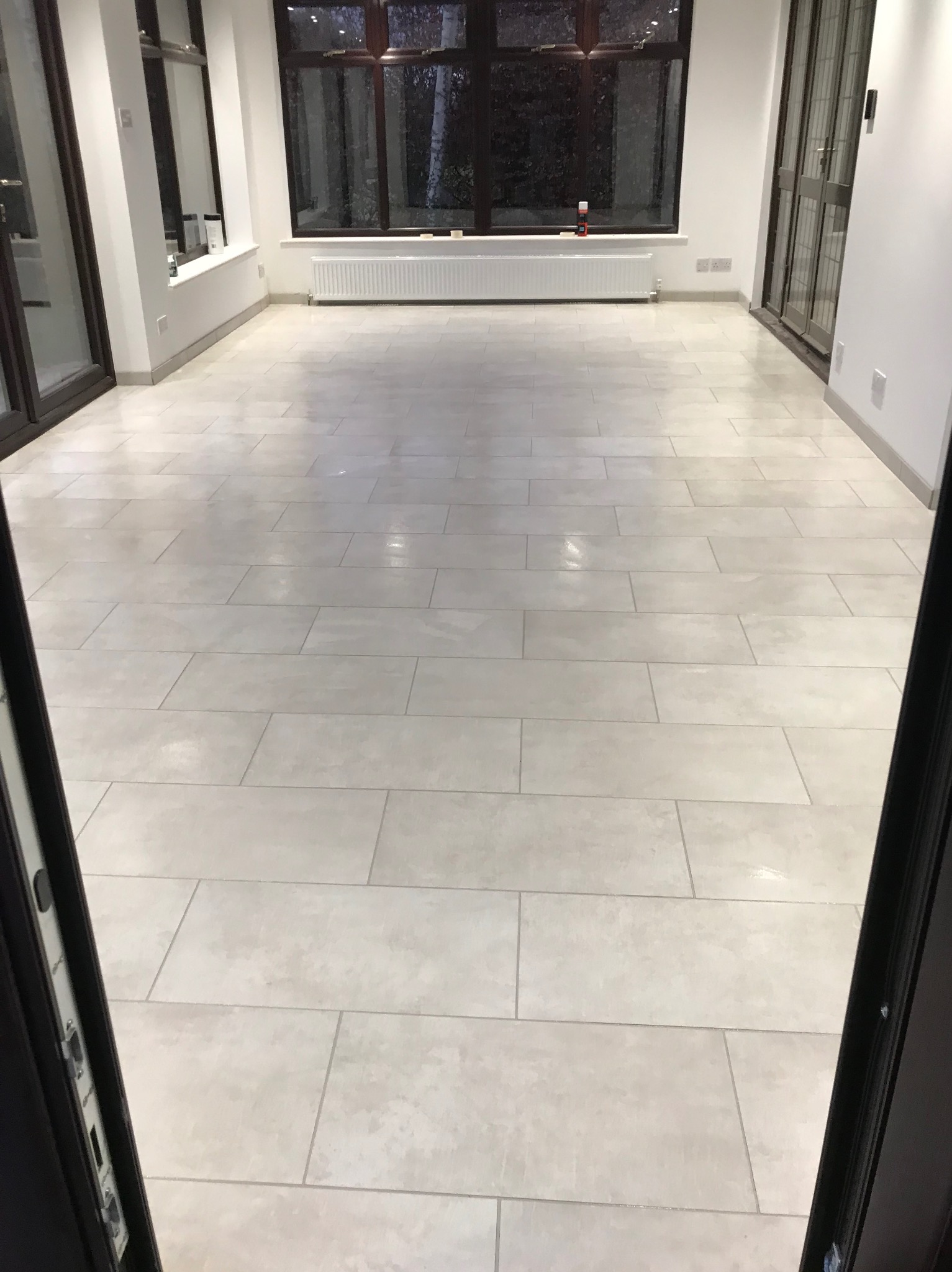 Jasna, prostokątna podłoga z płytek w nowocześnie wykończonym wnętrzu z dużymi oknami i grzejnikiem. Widok zza otwartych drzwi.