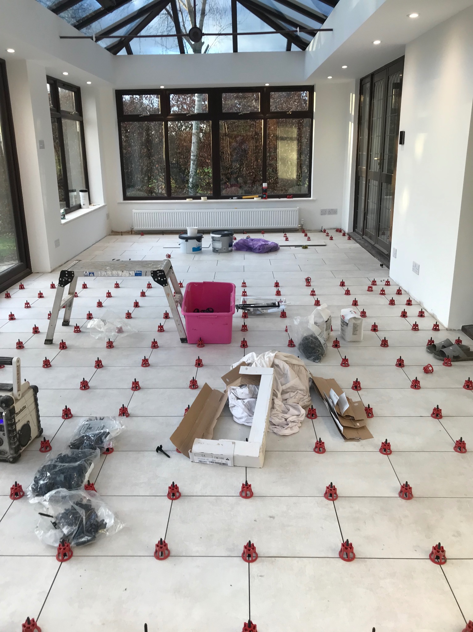 Jasne płytki podłogowe w oranżerii, wyrównane za pomocą czerwonych klipsów i czarnych klinów. Widoczna drabina, radio i akcesoria do układania płytek.