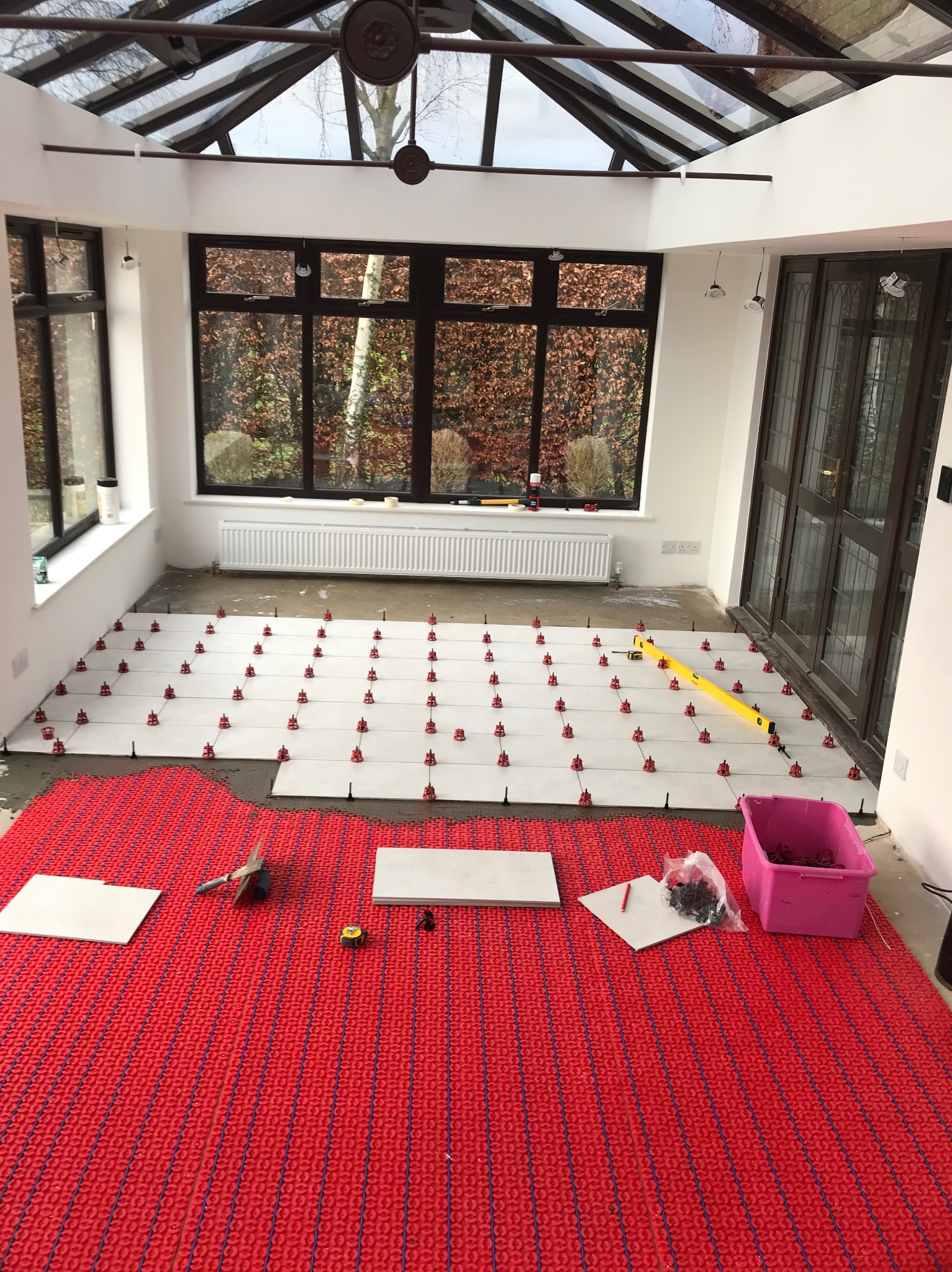 Układanie białych płytek na czerwonej macie grzewczej w jasnym pomieszczeniu z dużymi oknami. Widoczne narzędzia i poziomica. Prace wykończeniowe w trakcie.