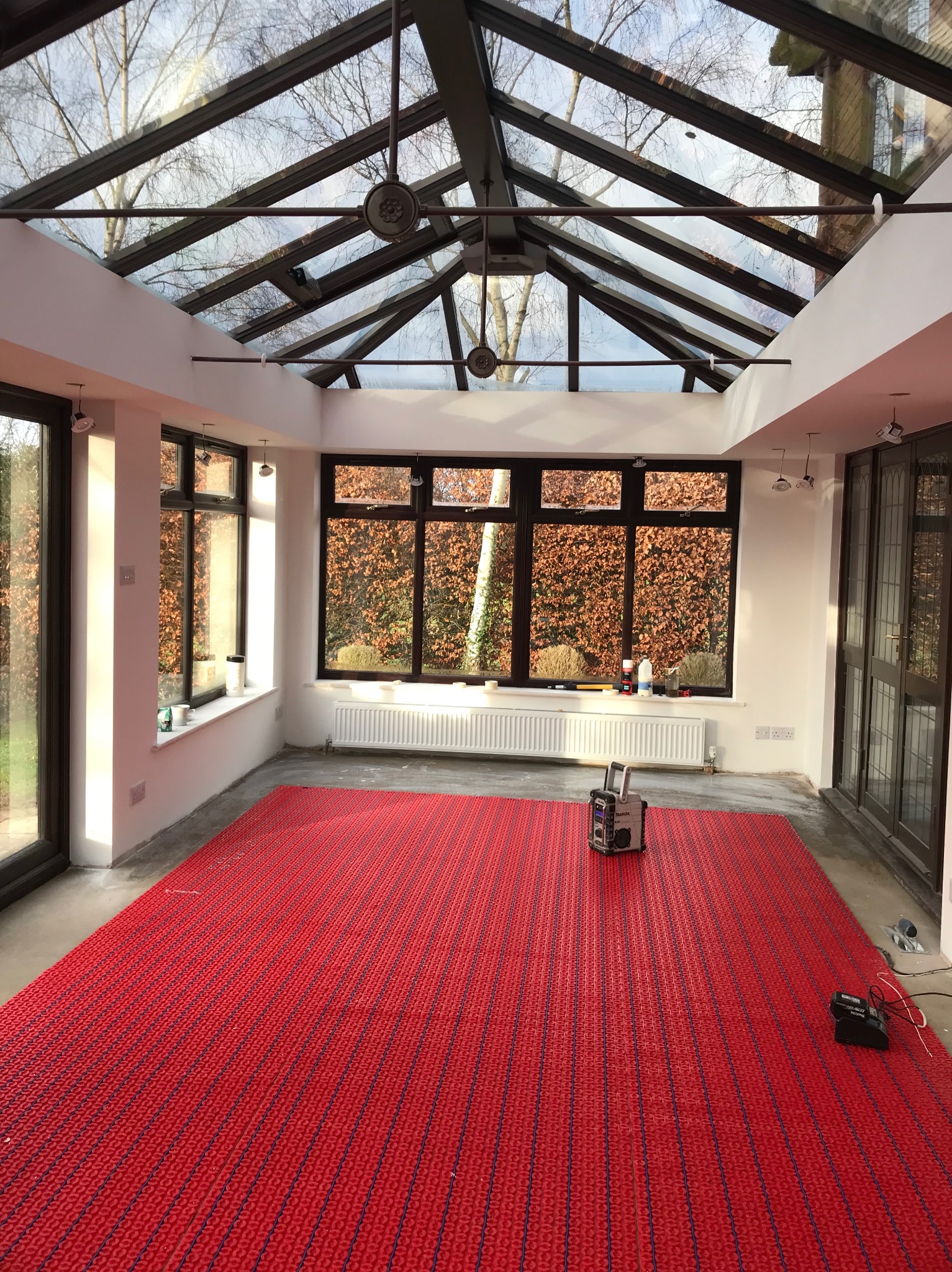 Wnętrze oranżerii z przeszklonym dachem i czerwonym systemem ogrzewania podłogowego. Widoczne okna z widokiem na zewnątrz i narzędzia pracy.