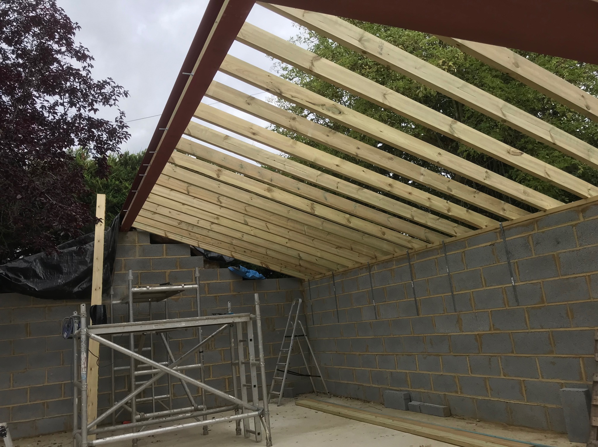 Budowa dachu z drewnianych belek na murowanym budynku. Widoczne rusztowania i drabina. Konstrukcja w trakcie realizacji, z widocznymi elementami mocującymi.