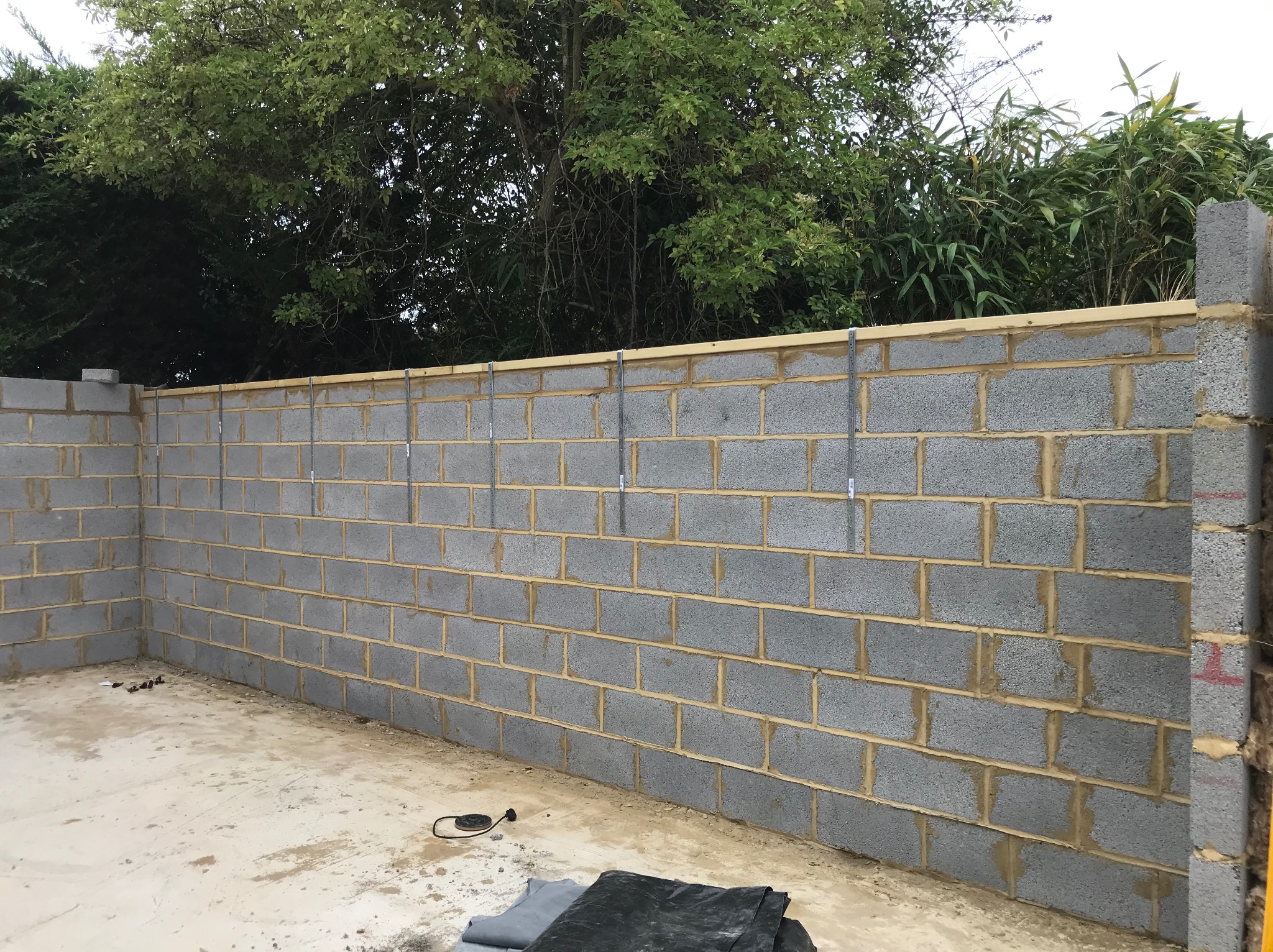 Ściana z szarych bloczków betonowych, z widocznymi spoinami i metalowymi wspornikami, w trakcie budowy. W tle zieleń drzew i krzewów.