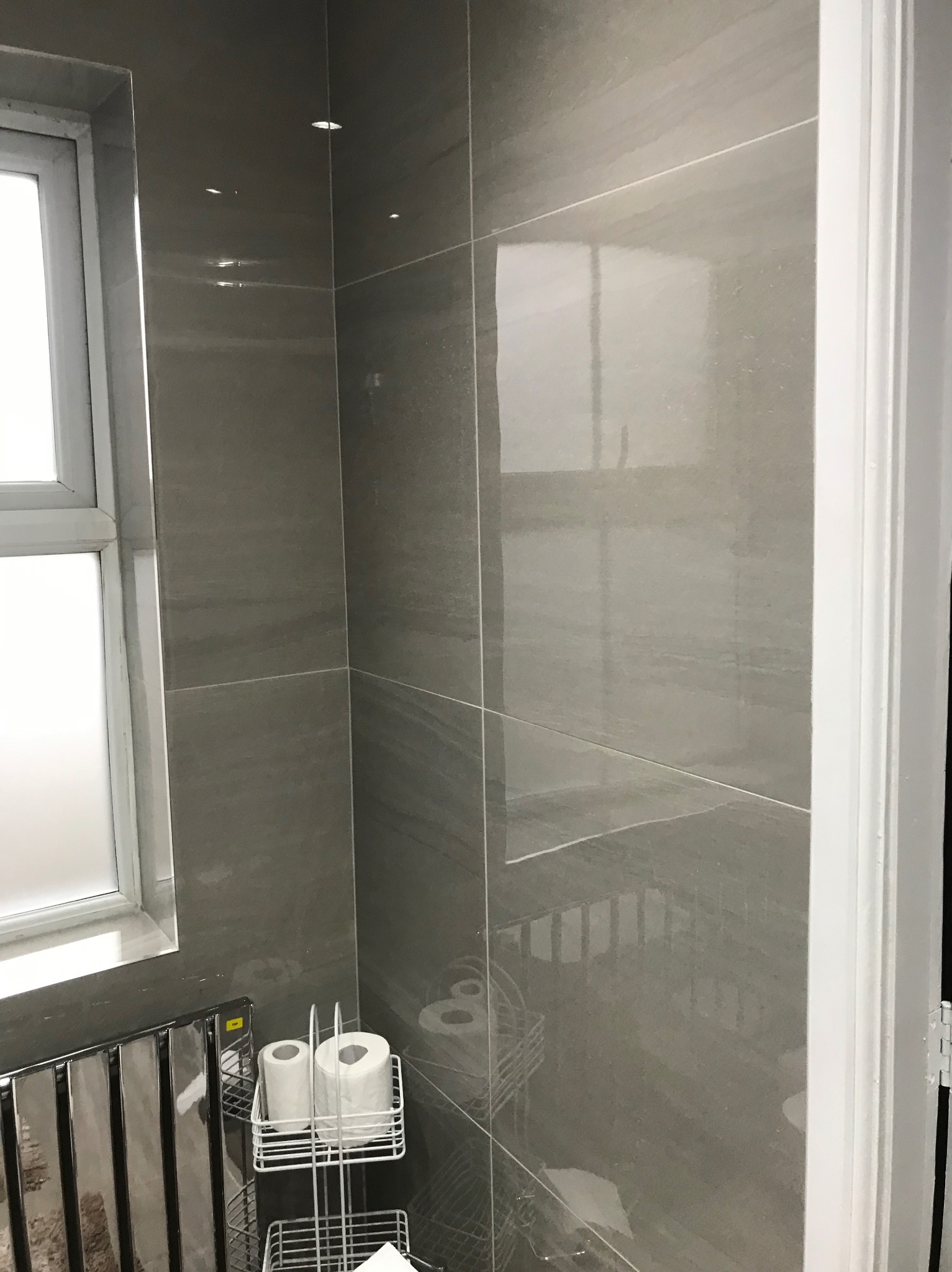 Wykończona ściana w łazience z szarymi, błyszczącymi płytkami imitującymi kamień, widoczny fragment okna i grzejnika drabinkowego z akcesoriami.