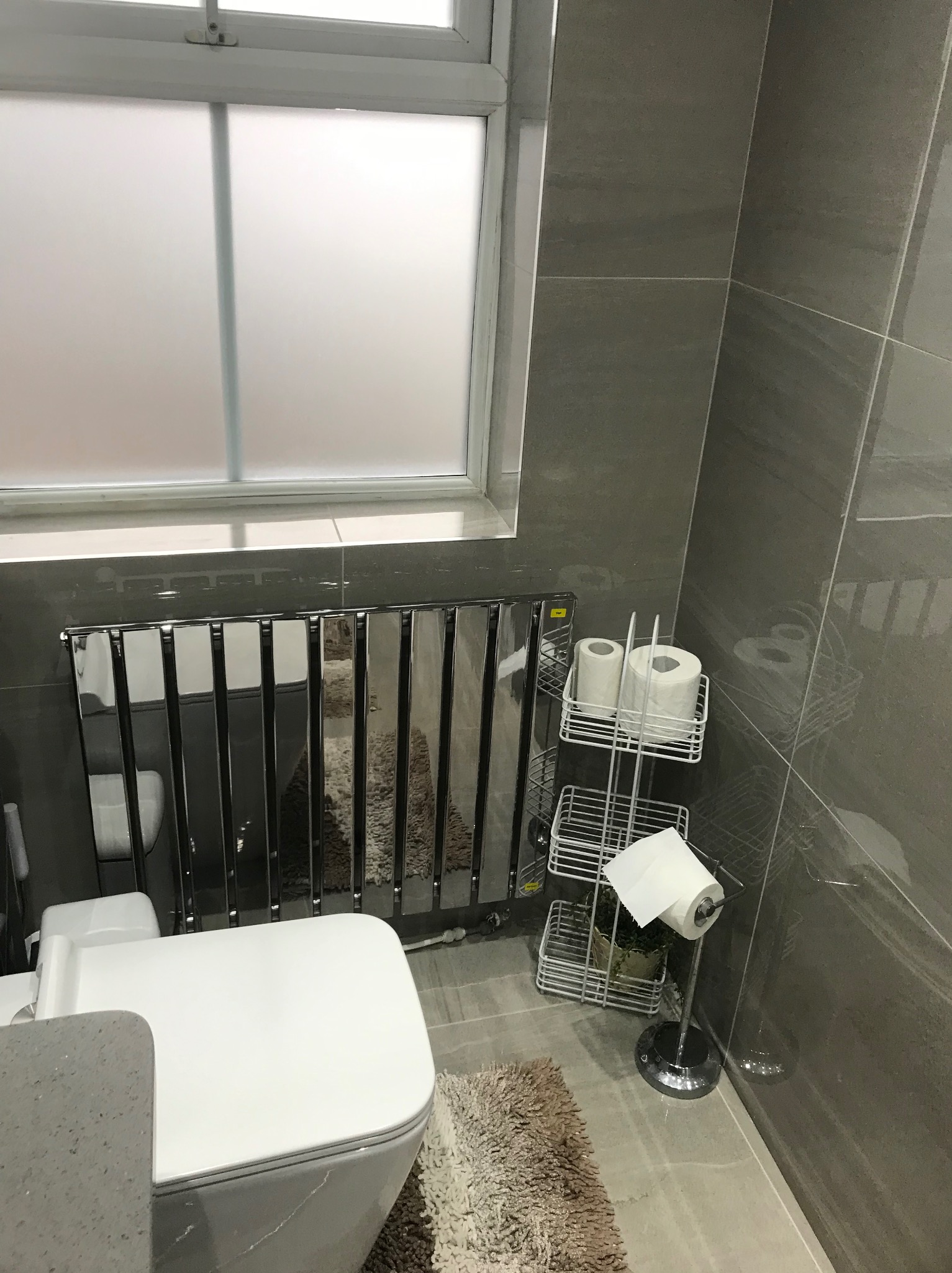 Nowoczesna łazienka z szarymi płytkami, chromowanym grzejnikiem drabinkowym i toaletą. Widoczny stojak na papier toaletowy i okno z matową szybą.