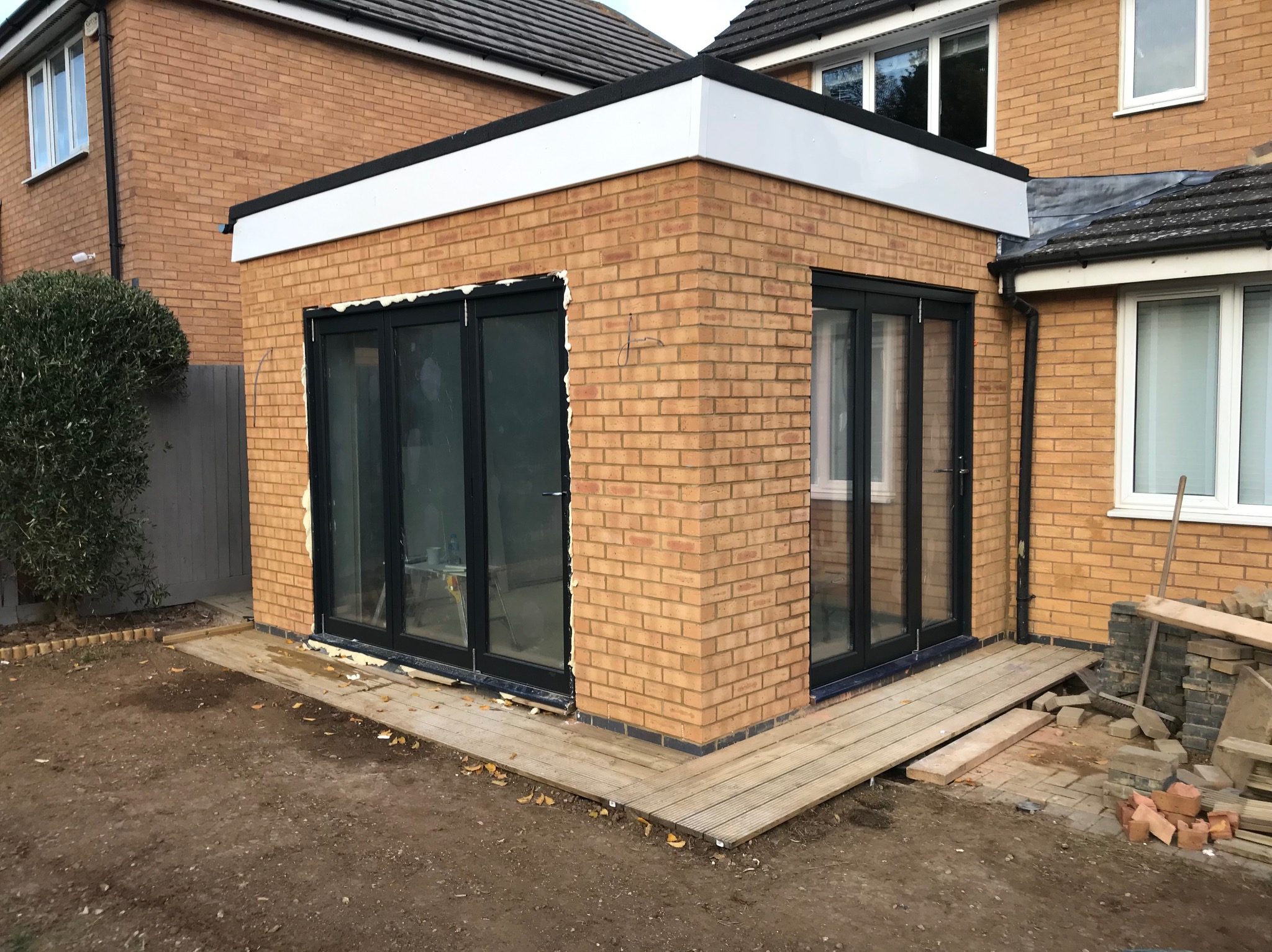 Dobudowa do domu z cegły klinkierowej z czarnymi, przeszklonymi drzwiami balkonowymi i białym wykończeniem dachu. Widoczne elementy drewnianego tarasu.