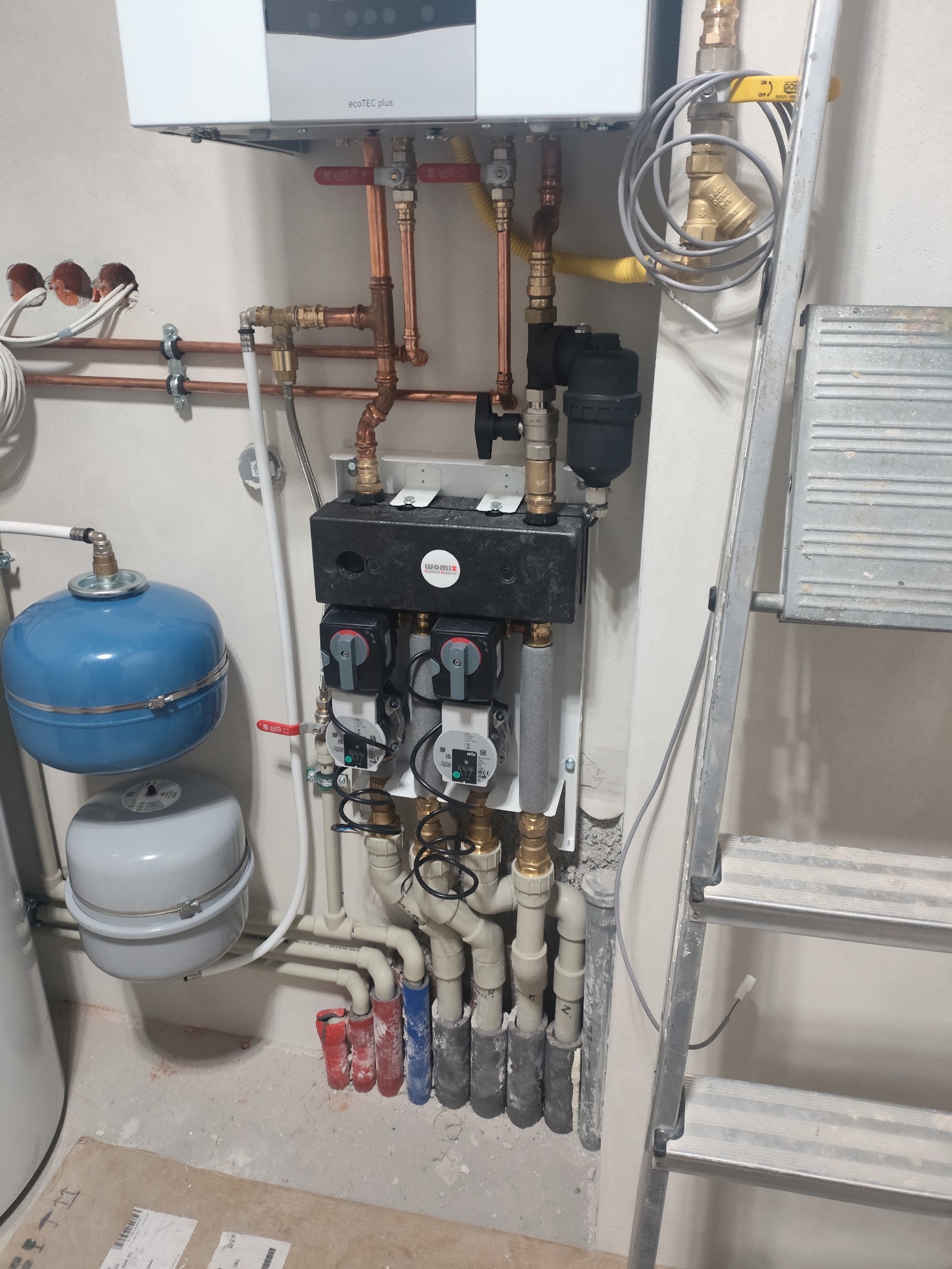 Złożona instalacja grzewcza z kotłem gazowym, pompami obiegowymi i zbiornikami wyrównawczymi. Widoczne miedziane rury, zawory i drabina oparta o ścianę.