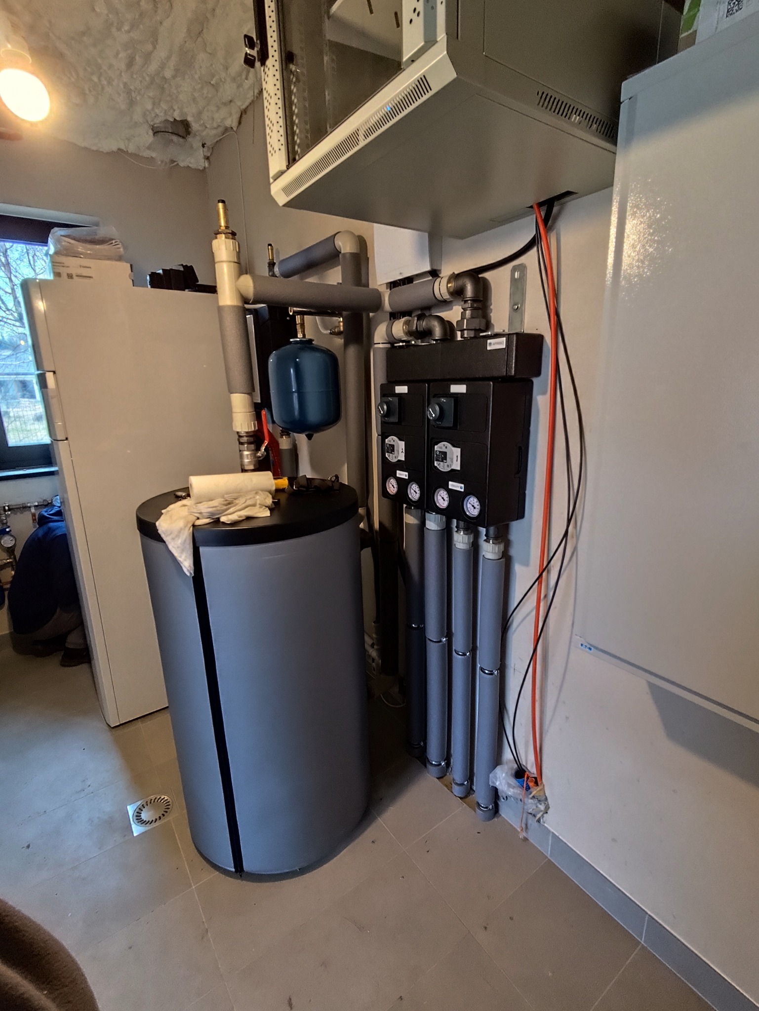 Szary zbiornik akumulacyjny z niebieskim naczyniem wzbiorczym i czarną szafką sterowniczą z manometrami, rurami i przewodami w kotłowni. Widoczny fragment okna.