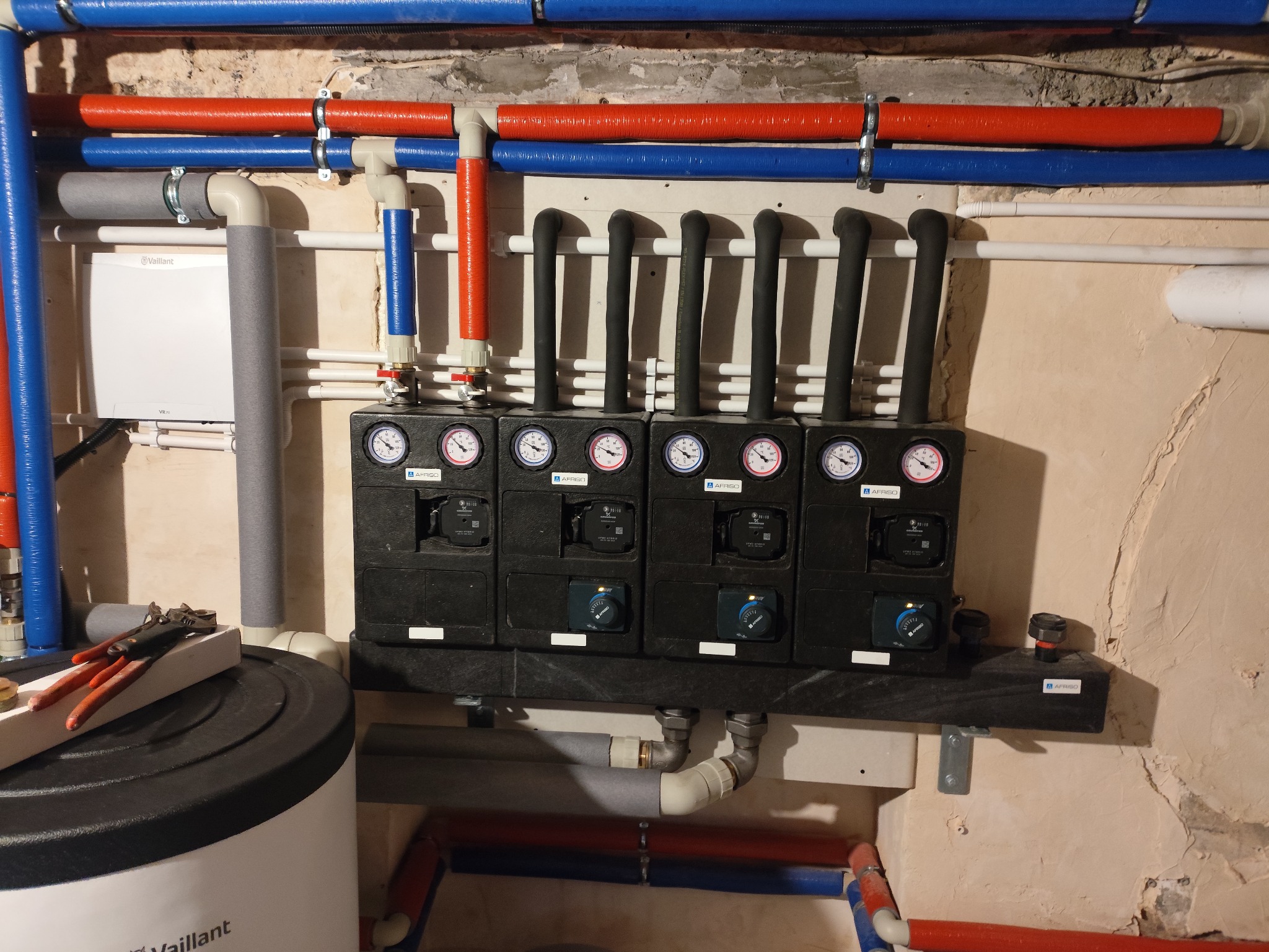 Rozdzielacz ogrzewania podłogowego z rurami w kolorach niebieskim i czerwonym, manometry, pompy obiegowe, zbiornik Vaillant i narzędzia hydrauliczne.