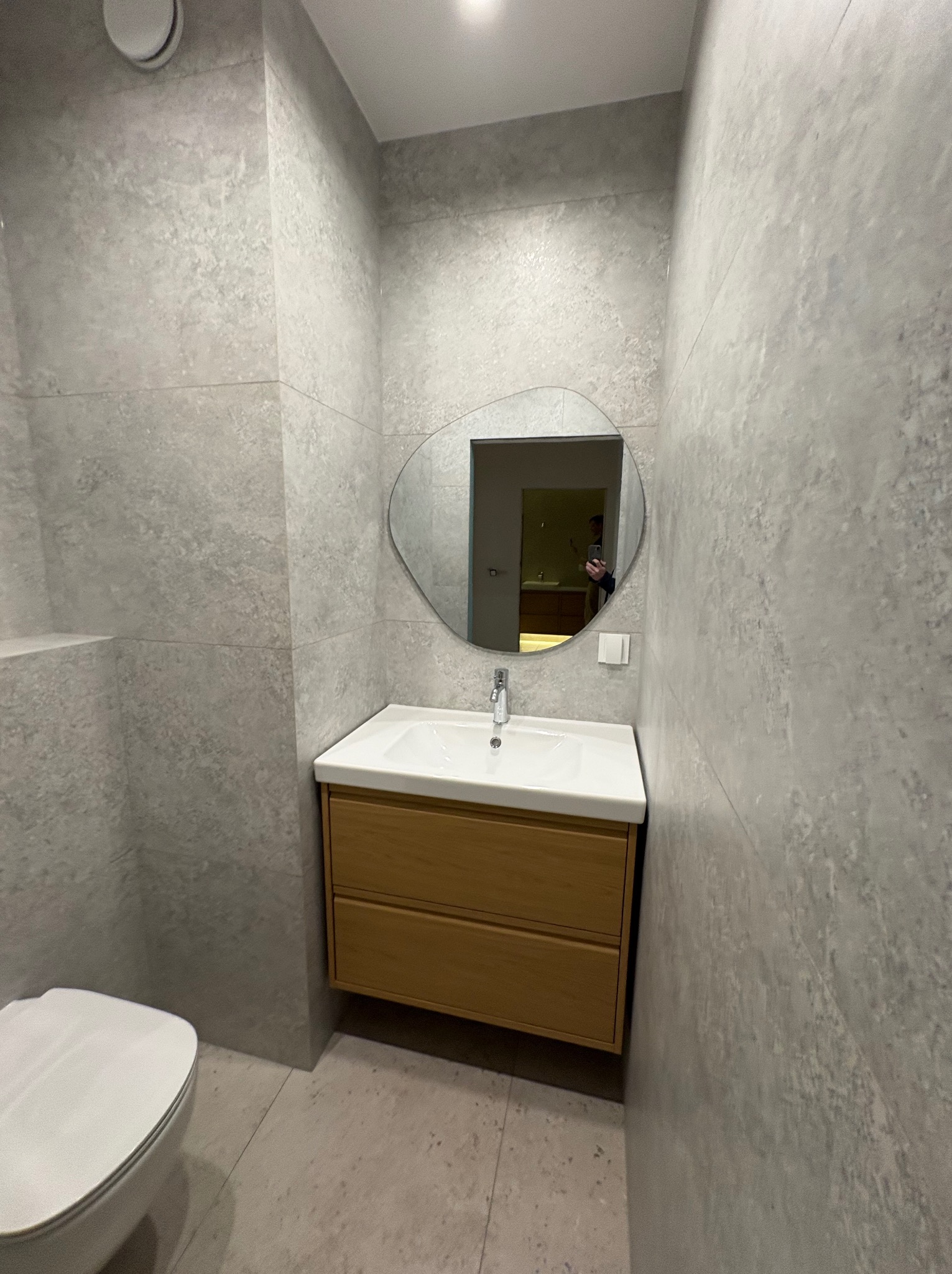 Wykończona łazienka w szarościach z drewnianą szafką pod umywalką i asymetrycznym lustrem. Widoczna toaleta i płytki imitujące kamień na ścianach i podłodze.