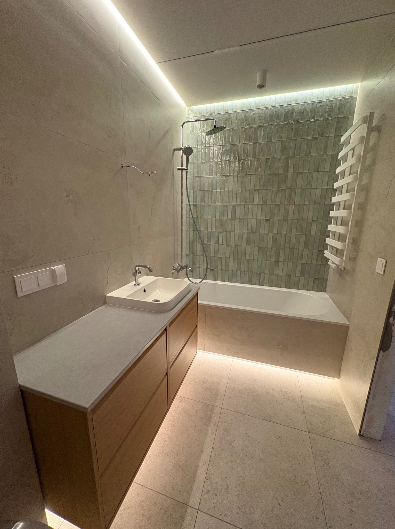 Nowoczesna łazienka z podświetlaną wanną i szafką pod umywalką. Beżowe płytki, zielona ściana nad wanną, grzejnik drabinkowy. Minimalistyczny design z akcentami świetlnymi.