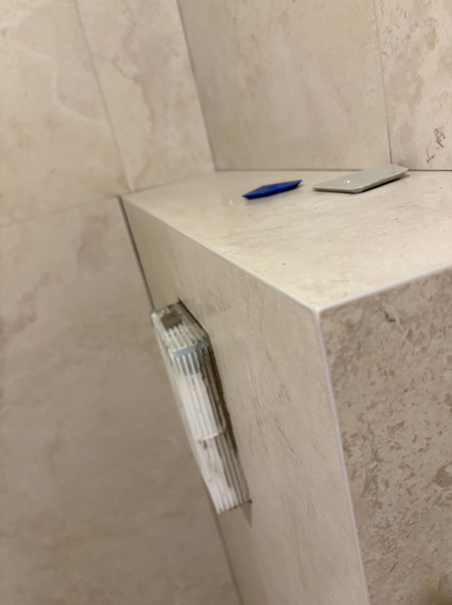 Półka w łazience wyłożona beżowymi płytkami, z narzędziami glazurniczymi. Widoczne fugi i listwa wykończeniowa. Detal wykończenia wnętrza.